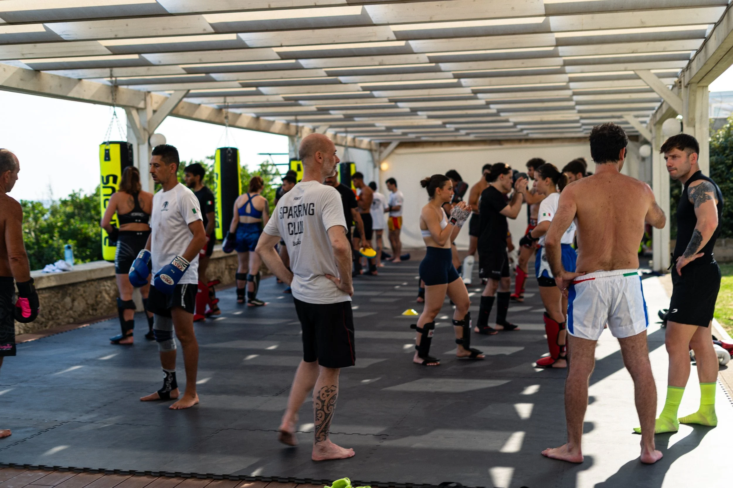 People participating in a mixed martial arts or boxing training session outdoors under a pergola with wooden beams and sunlight filtering through, wearing athletic clothing and gloves.
