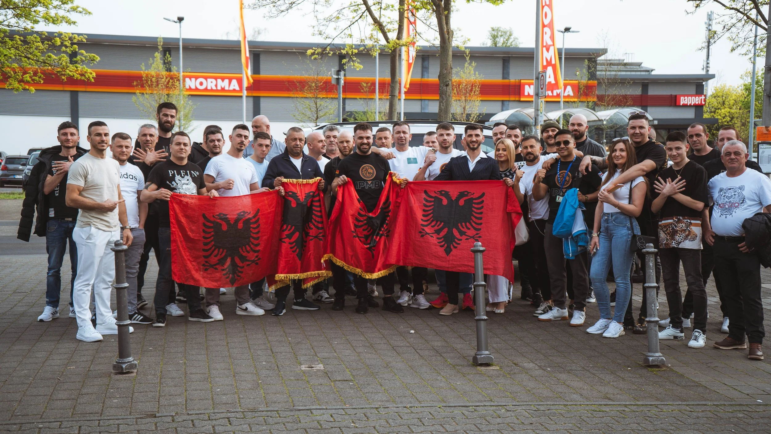Group of people posing outdoors, holding Albanian flags, standing in front of a building with red and orange signage, some making gestures or pointing.