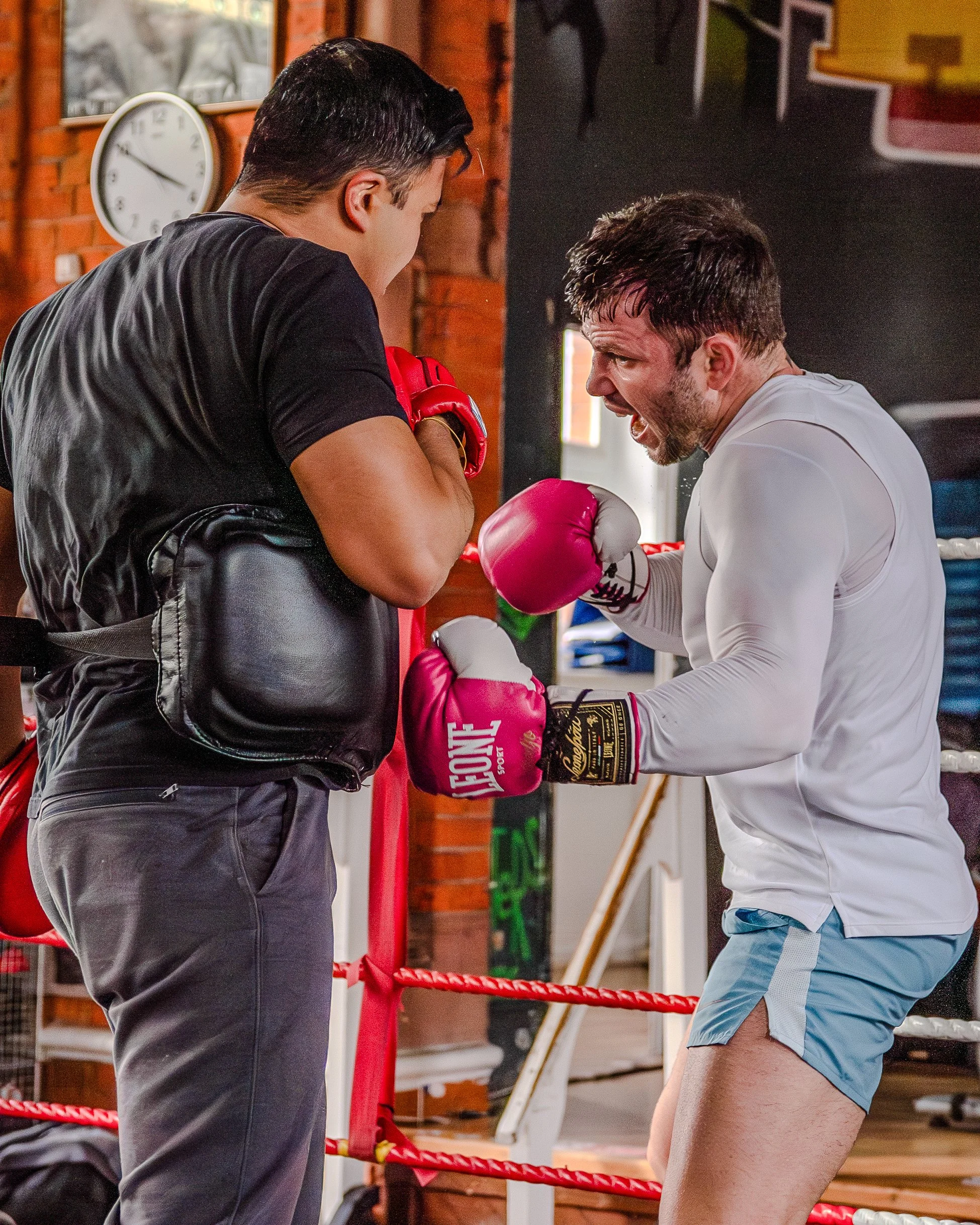 Two men engaged in a boxing training session in a gym, one man wearing red boxing gloves and black attire, and the other man in a white shirt and light blue shorts, both inside a boxing ring with red ropes.