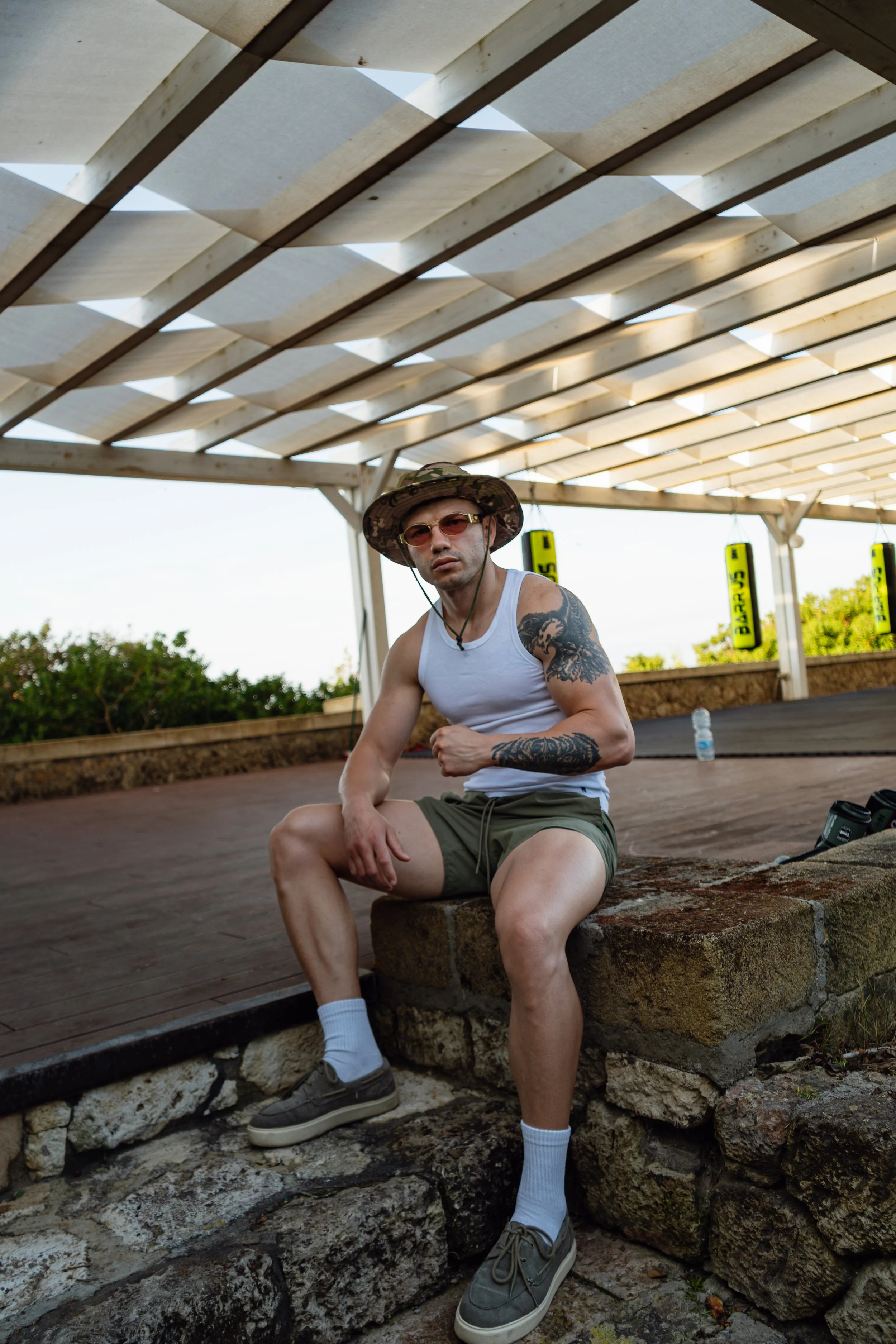 A man sitting on stone steps outdoors beneath a wooden pergola, wearing sunglasses, a wide-brimmed hat, a white tank top, shorts, and sneakers, with a water bottle nearby and Bag tags hanging in the background.