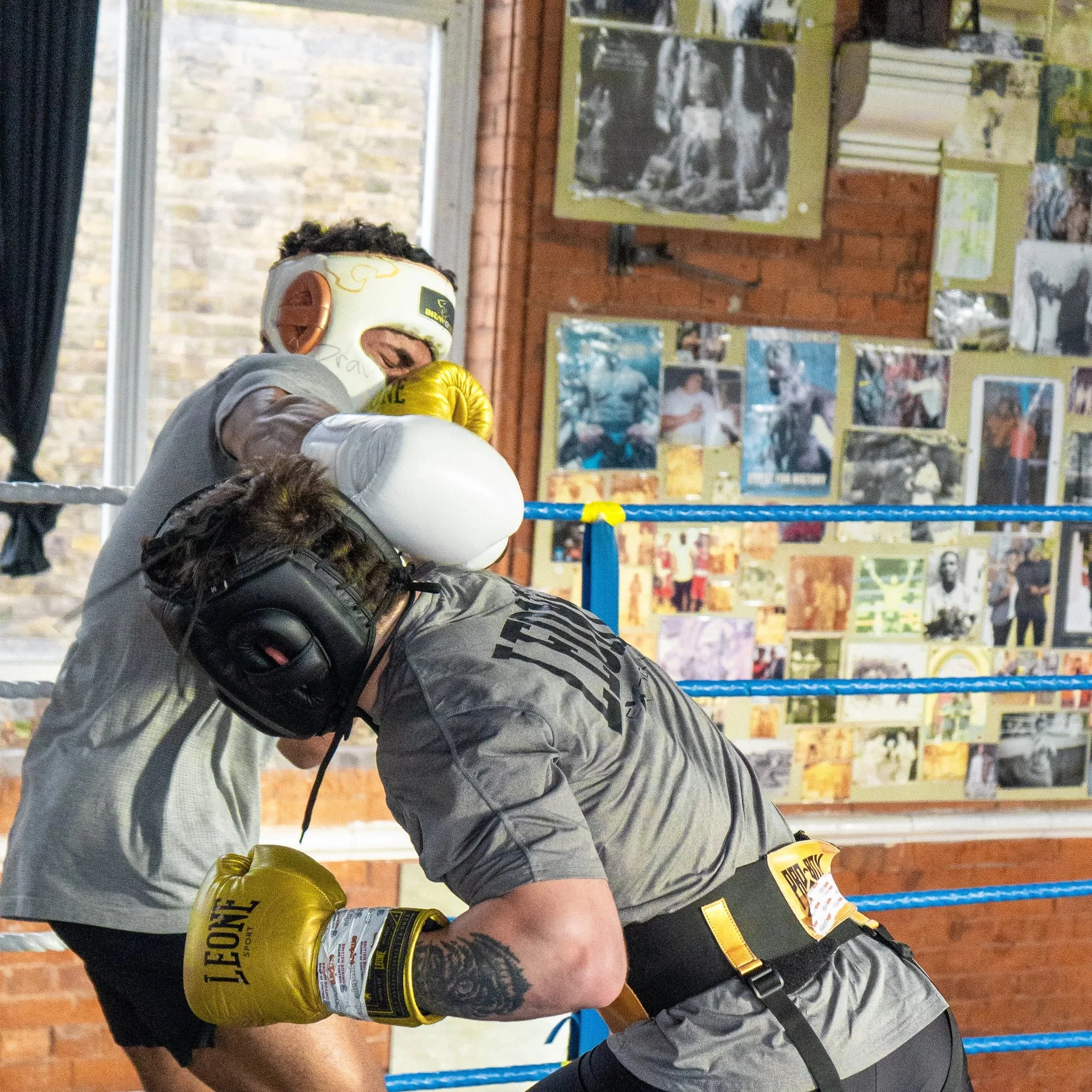 A young man wearing boxing gloves and headgear is training in a boxing gym, with photos and posters on the walls and a boxing ring in the background.