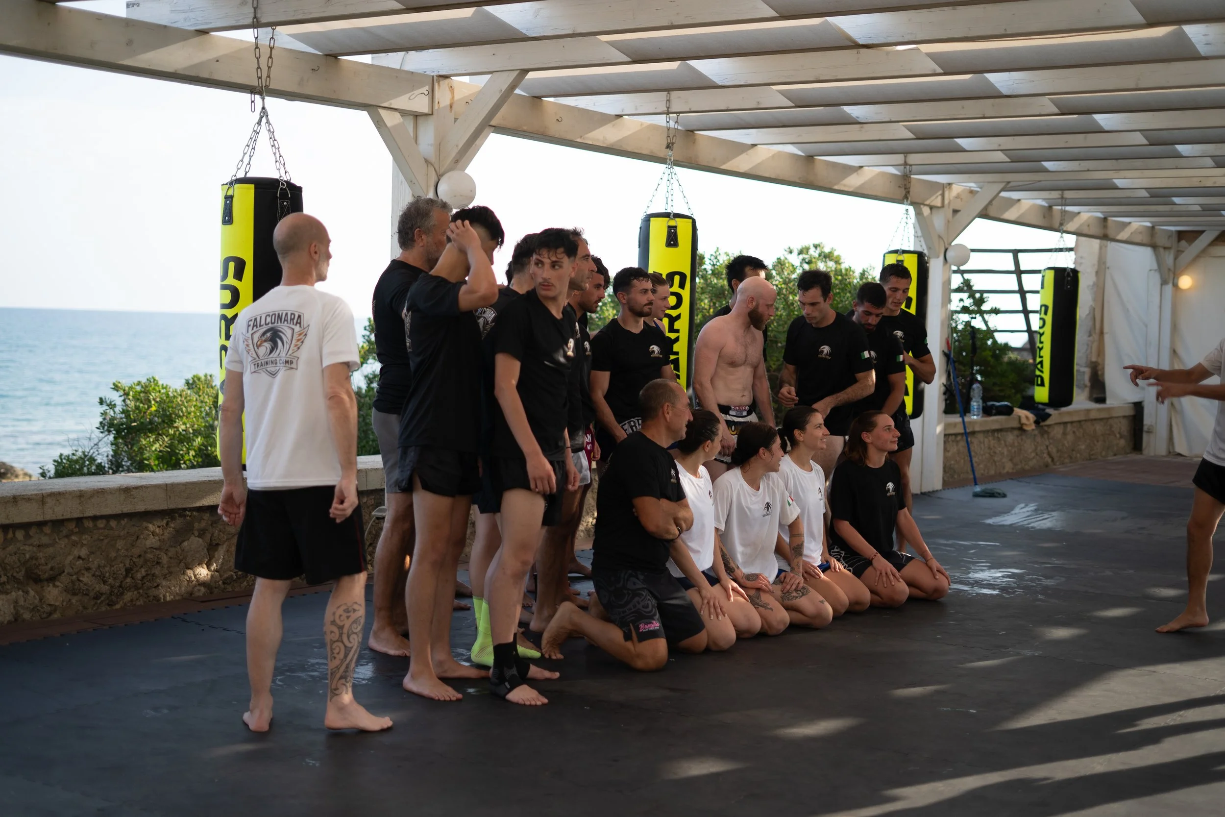 A group of people, some kneeling and some standing, are gathered under a canopy with hanging punching bags near a body of water. Some individuals are wearing athletic clothing including fight shorts and t-shirts, indicating they are at a martial arts or fitness training session.