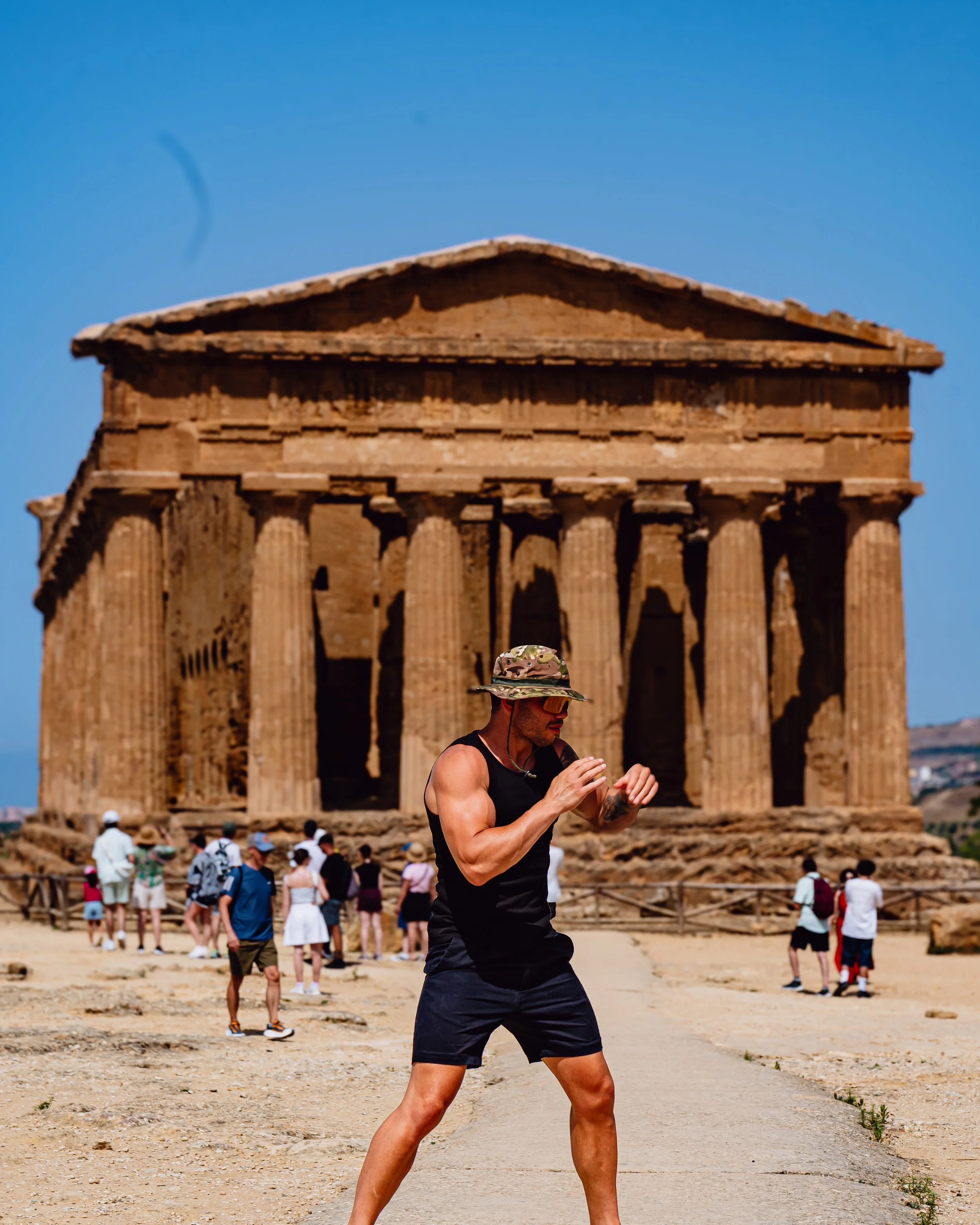 A man in a black tank top, shorts, and a camouflage hat appears to be exercising or practicing martial arts in front of the Parthenon in Athens, Greece during the daytime. There are many people in the background near the ancient ruins.