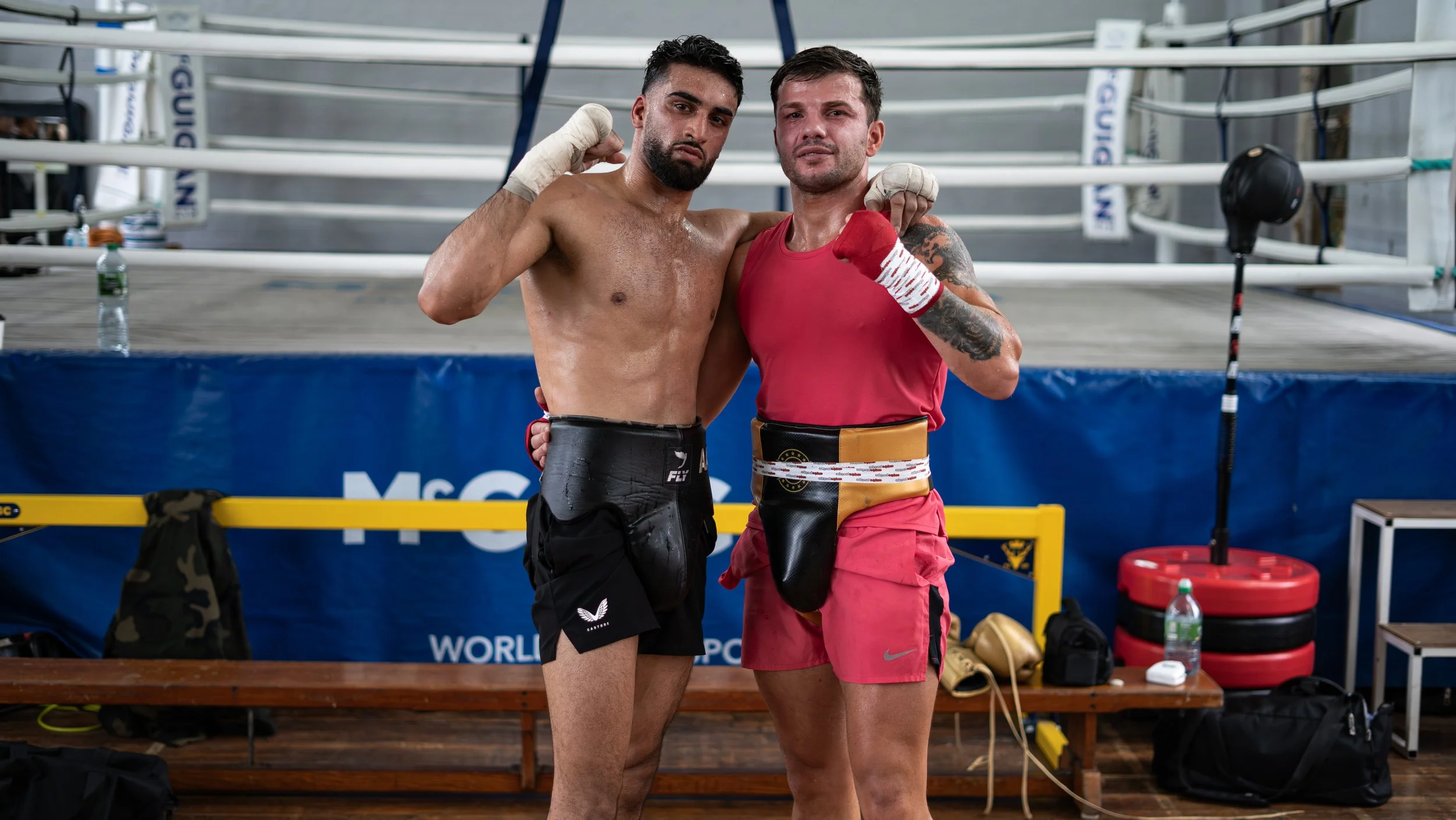 Two male fighters, one with a beard and the other with tattoos, pose together in a boxing ring after a match, raising their fists in victory and wearing boxing gloves.