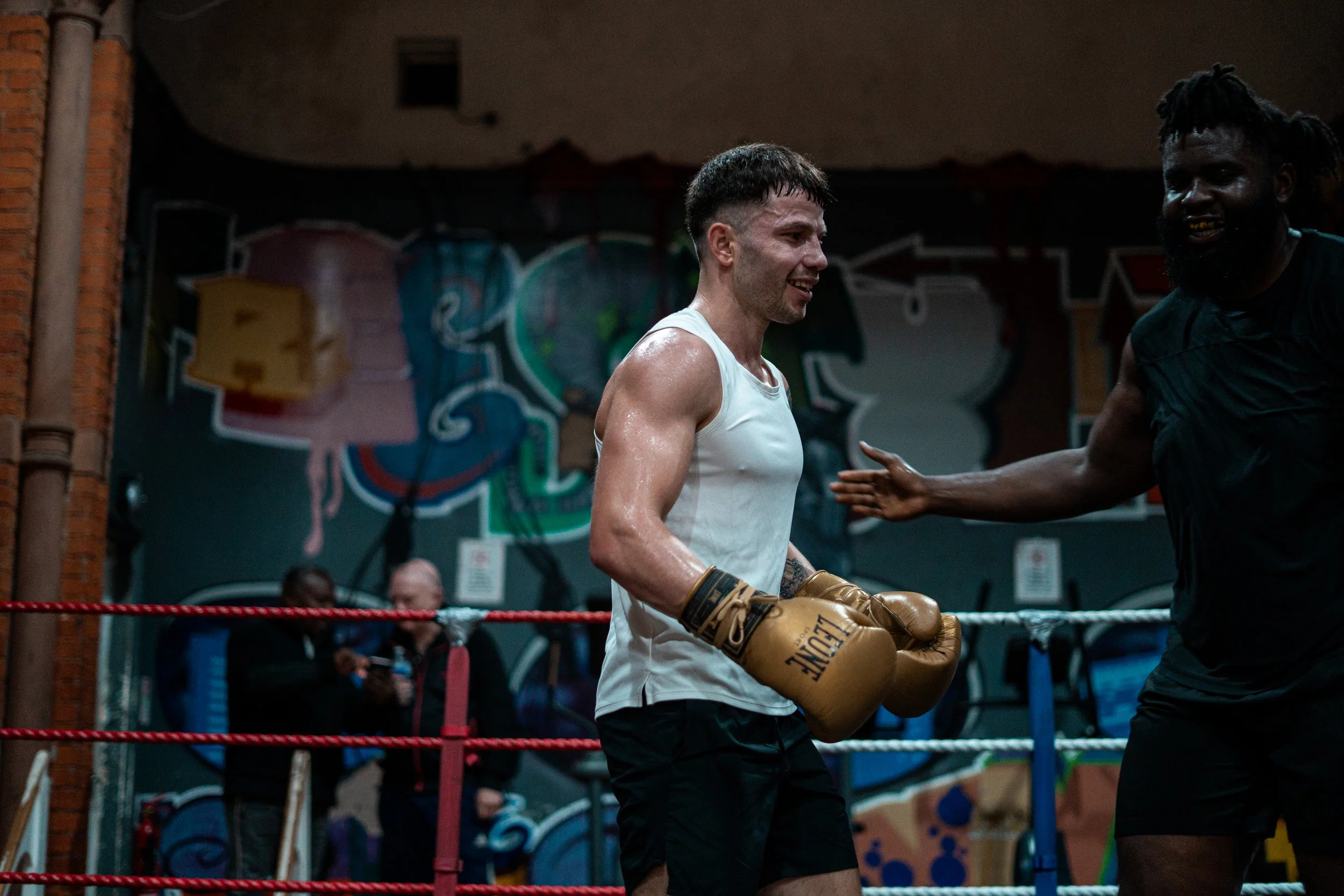 Two male boxers in a boxing ring after a match, smiling and appearing happy, with one wearing white and tan gloves and the other in dark clothing, against a background with colorful graffiti art.