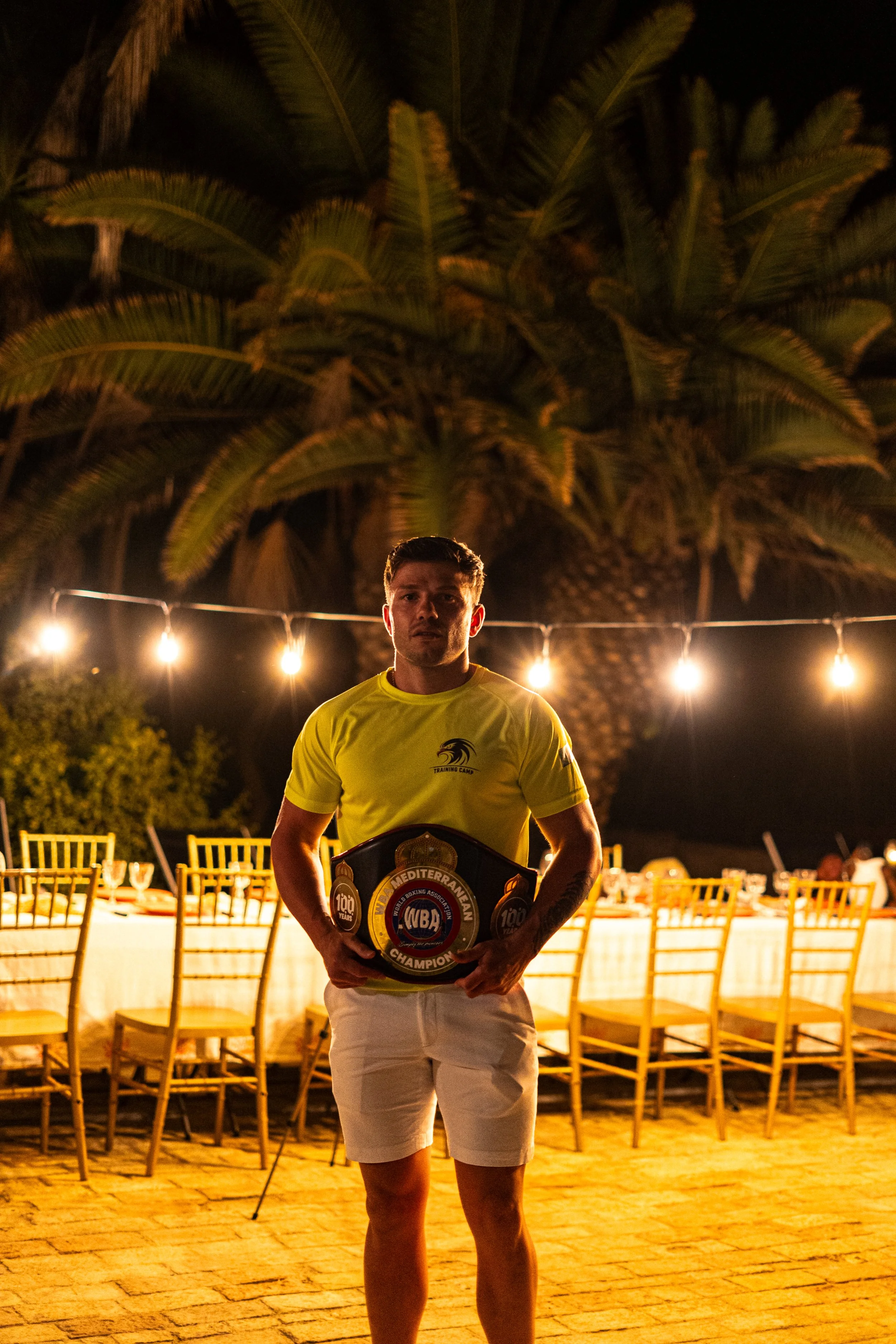 A man standing outdoors at night, holding a boxing championship belt, with a large palm tree and string lights in the background, seated at tables with yellow chairs in a festive setting.