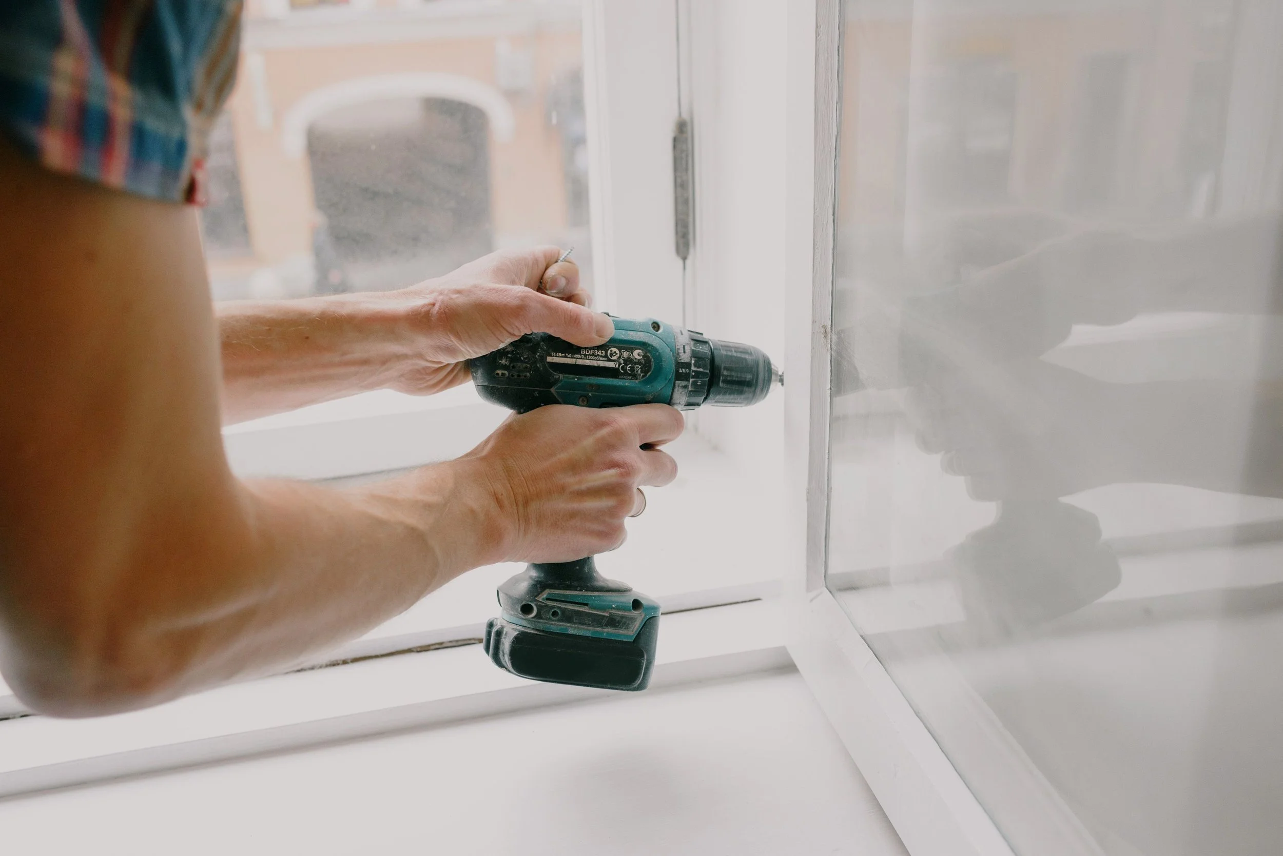 A person using a cordless drill to install or repair a white cabinet or furniture piece near a window.