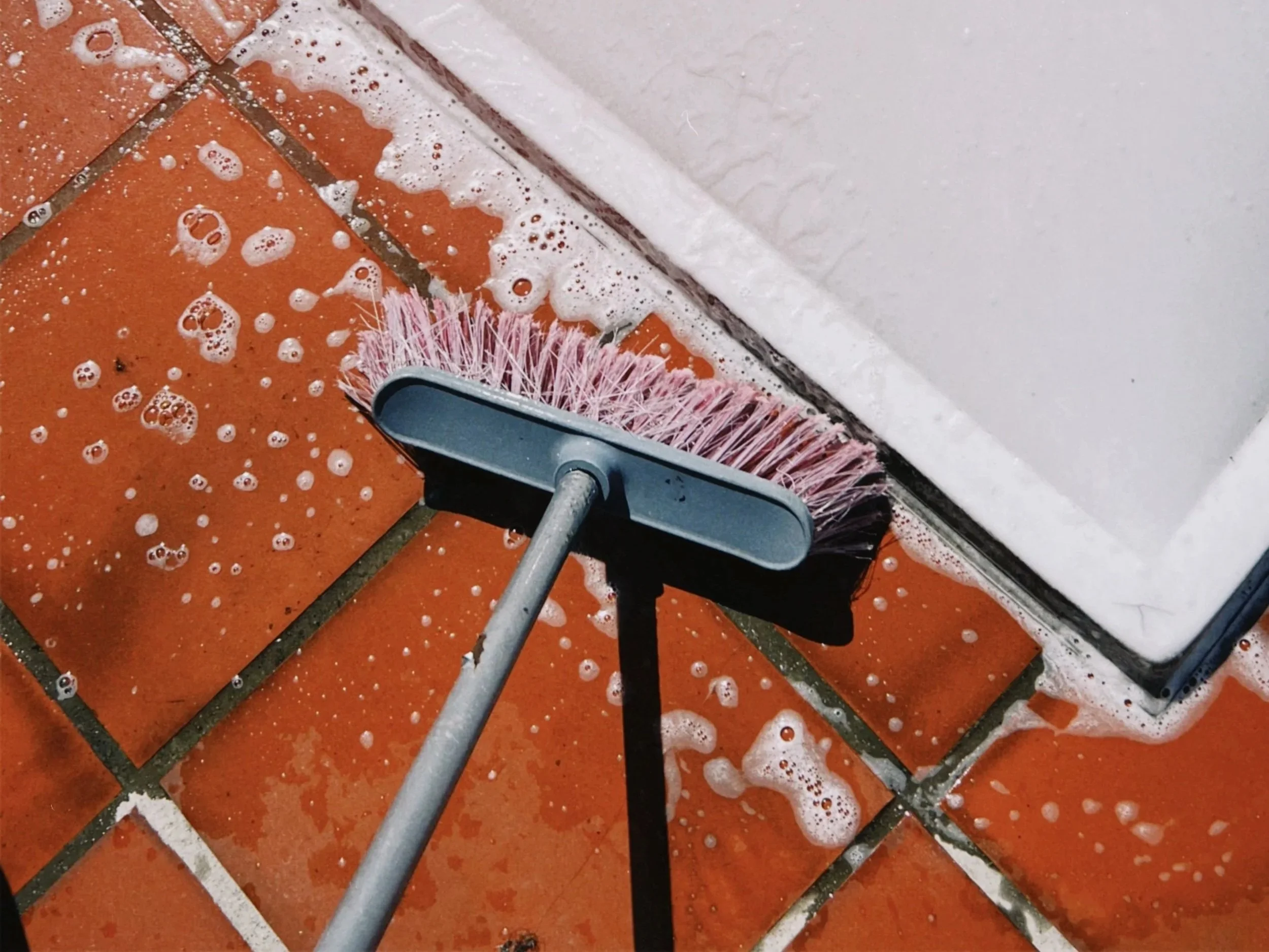 A brush cleaning a tiled red floor with soapy water
