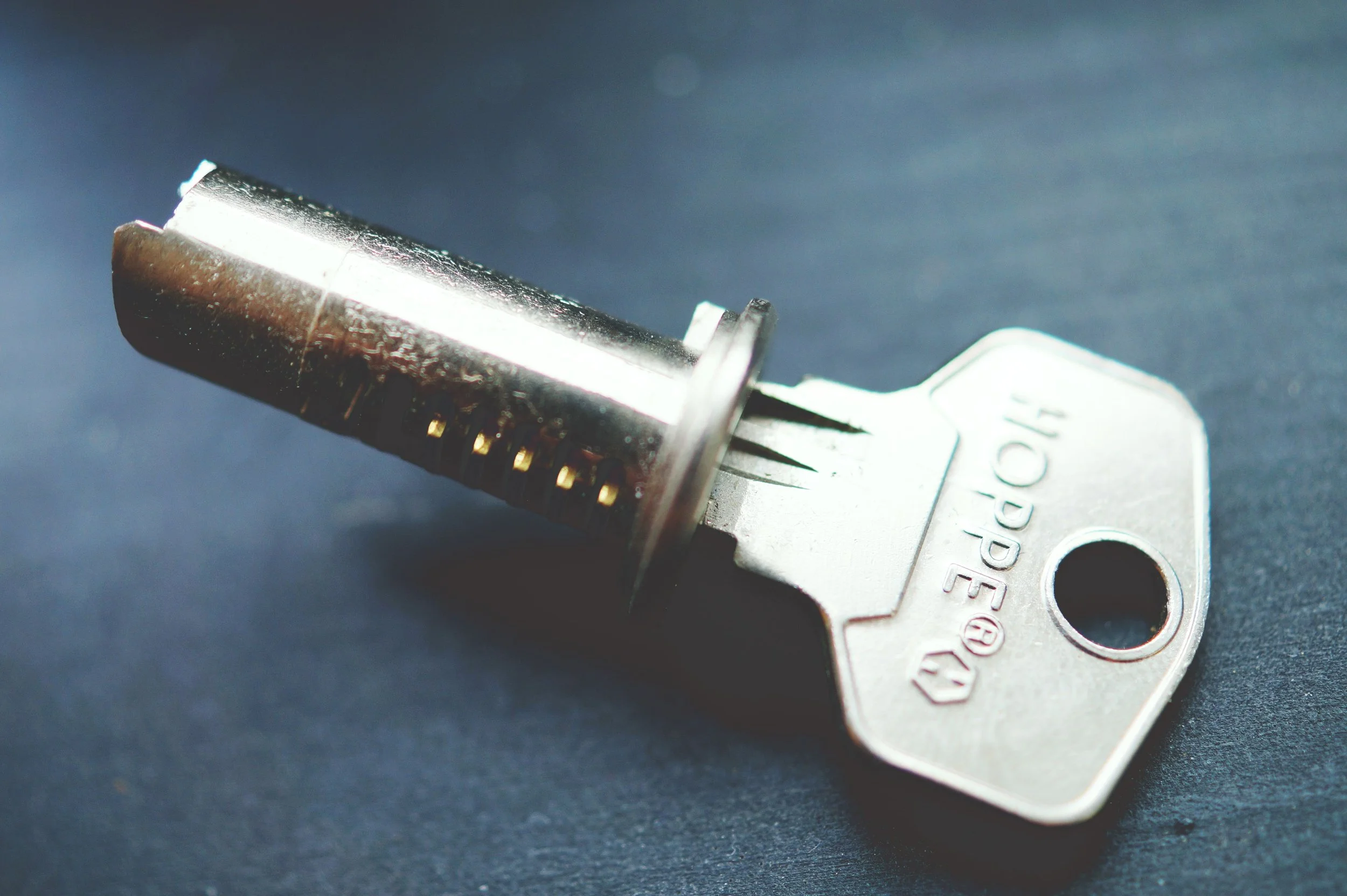 Close-up of a key with a metallic blade and a plastic head, lying on a dark surface. The key has a flat, silver-colored body with the word 'OPERA' engraved on it.