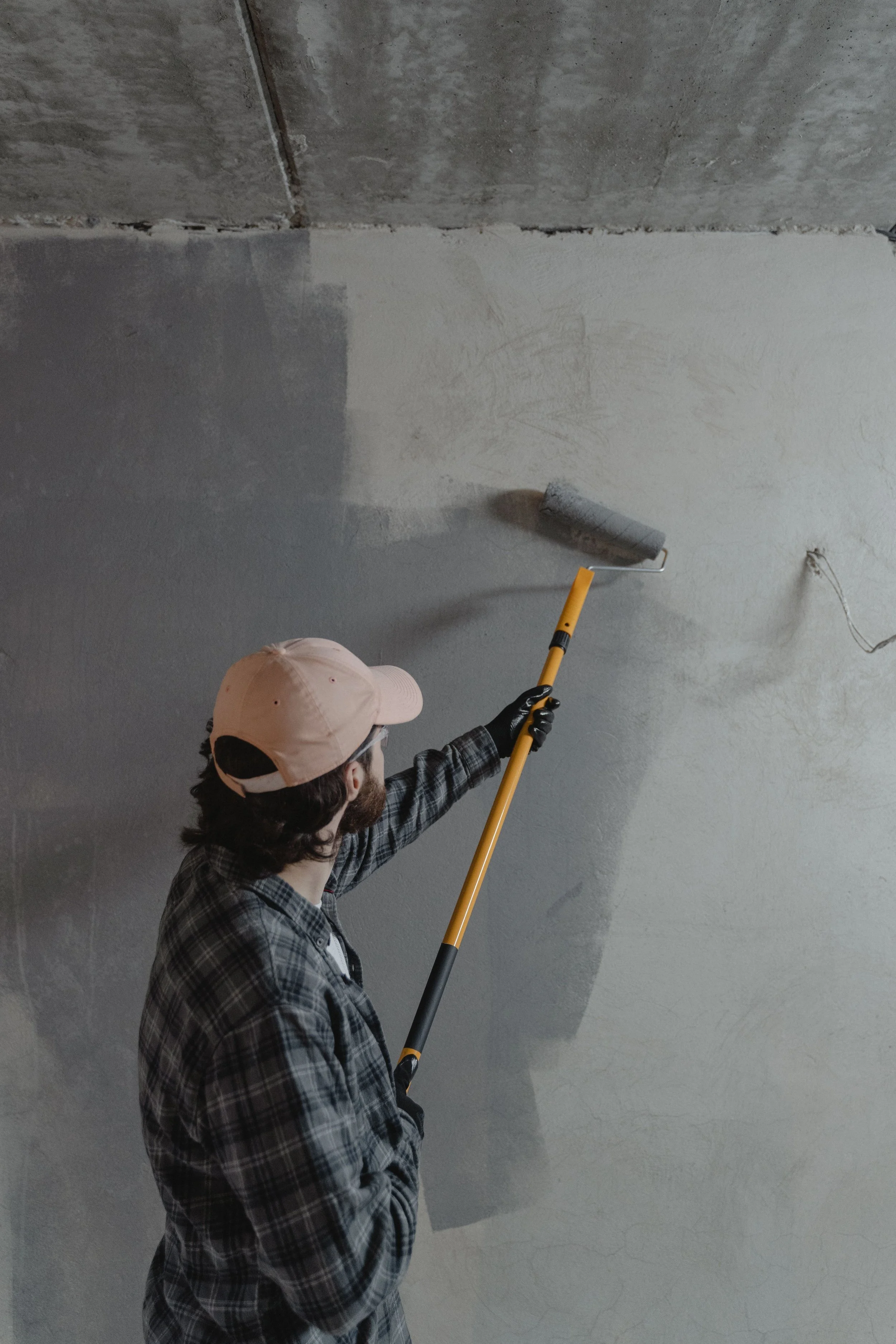 A person wearing a pink cap, glasses, and a plaid jacket painting a wall with gray paint using a paint roller.