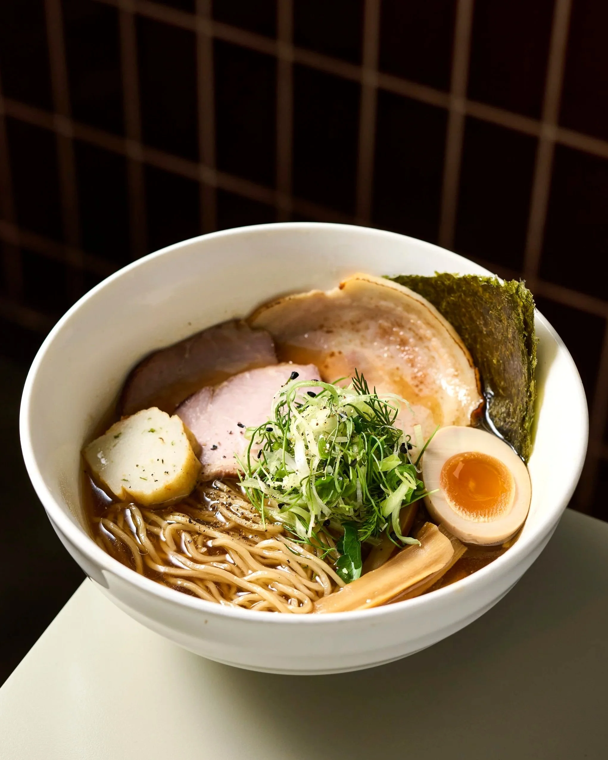 Close-up of a bowl of ramen noodles with half a soft-boiled egg, slices of pork, seaweed, green onions, and cilantro, set against a dark background.