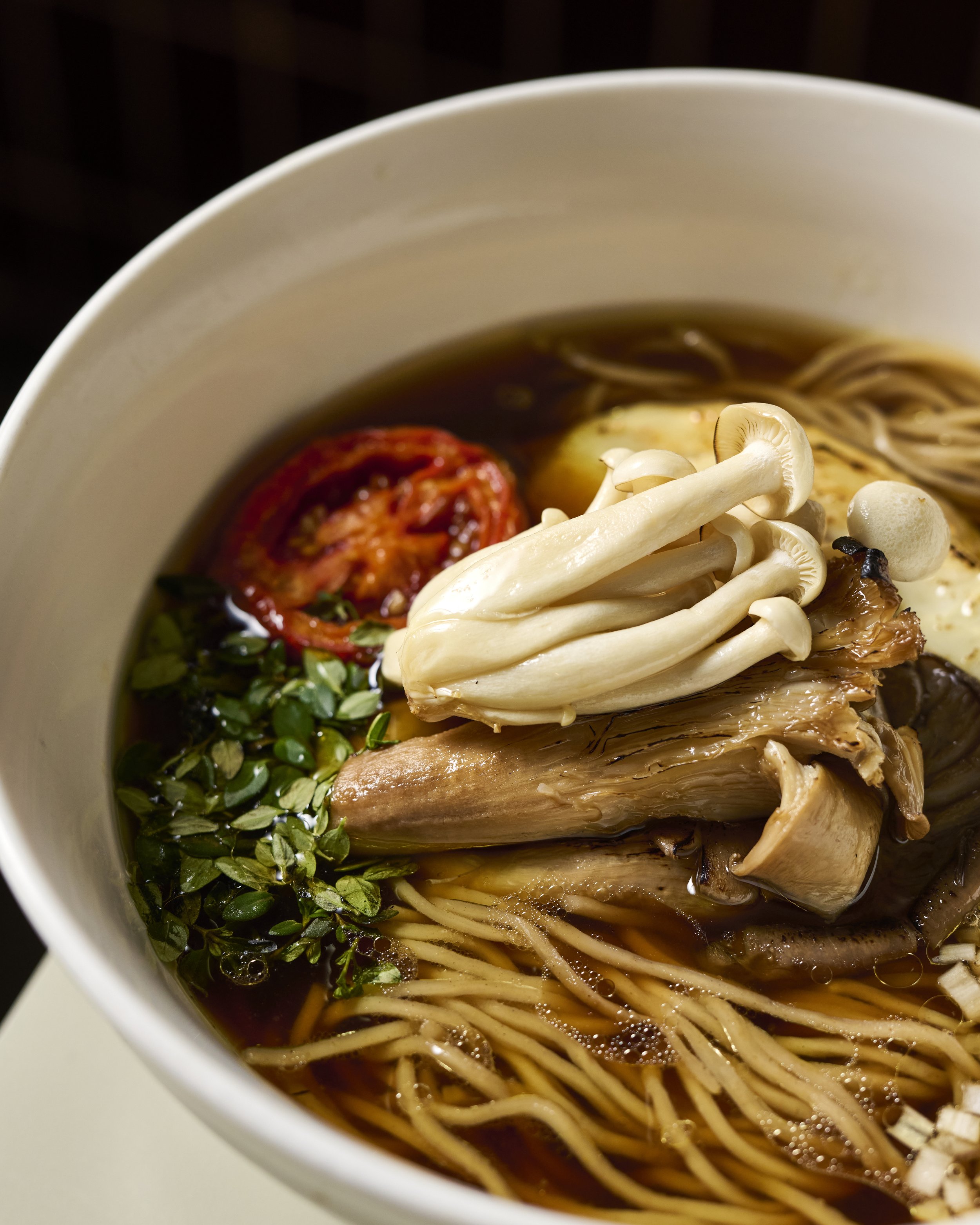 A ramen with a dark clear broth in a white bowl, topped with thyme leaves, enoki and shiitake mushroom, and folded noodles.