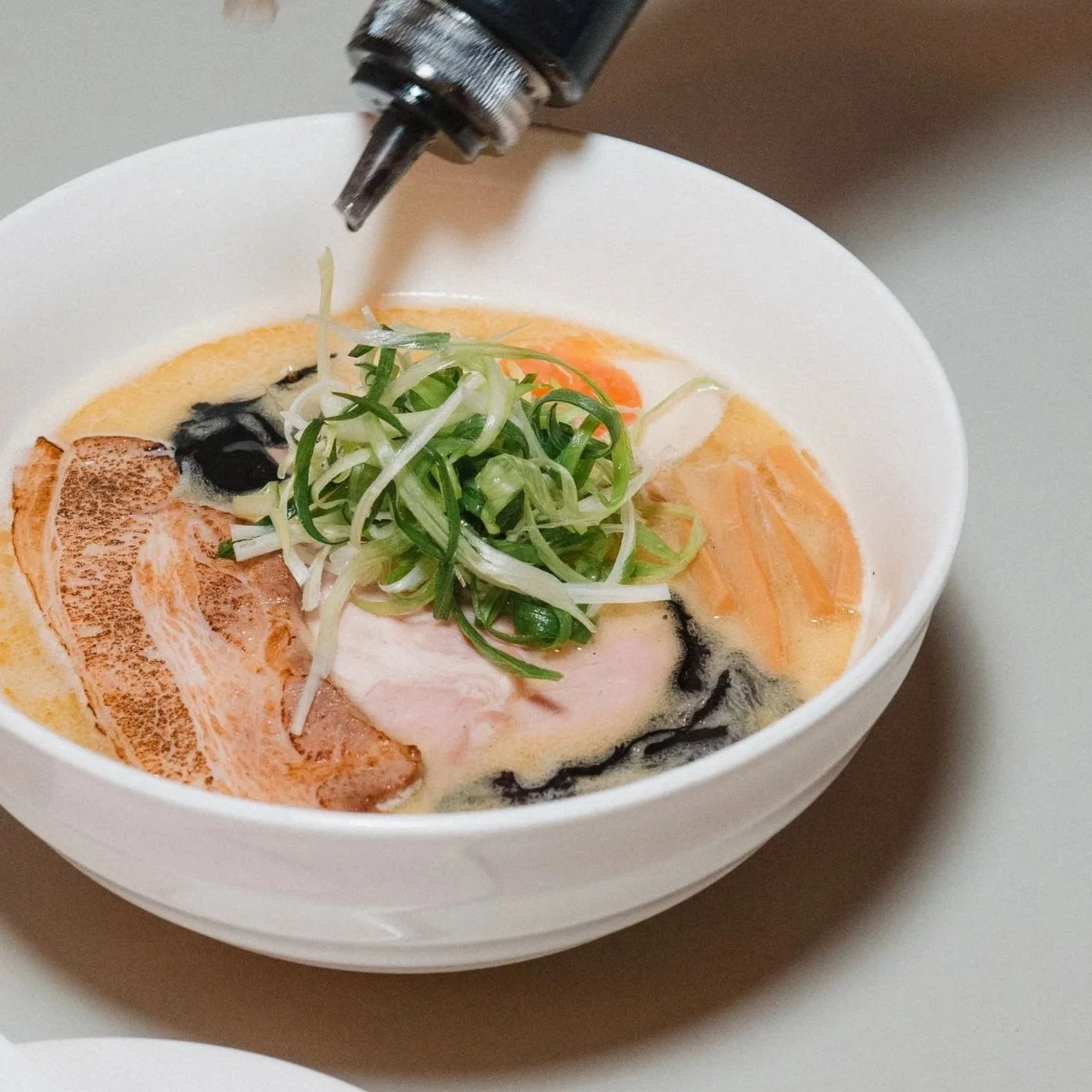 A white ramen bowl with a creamy soup inside and topped with pork and spring onion. A black garlic drizzle is being poured on top.