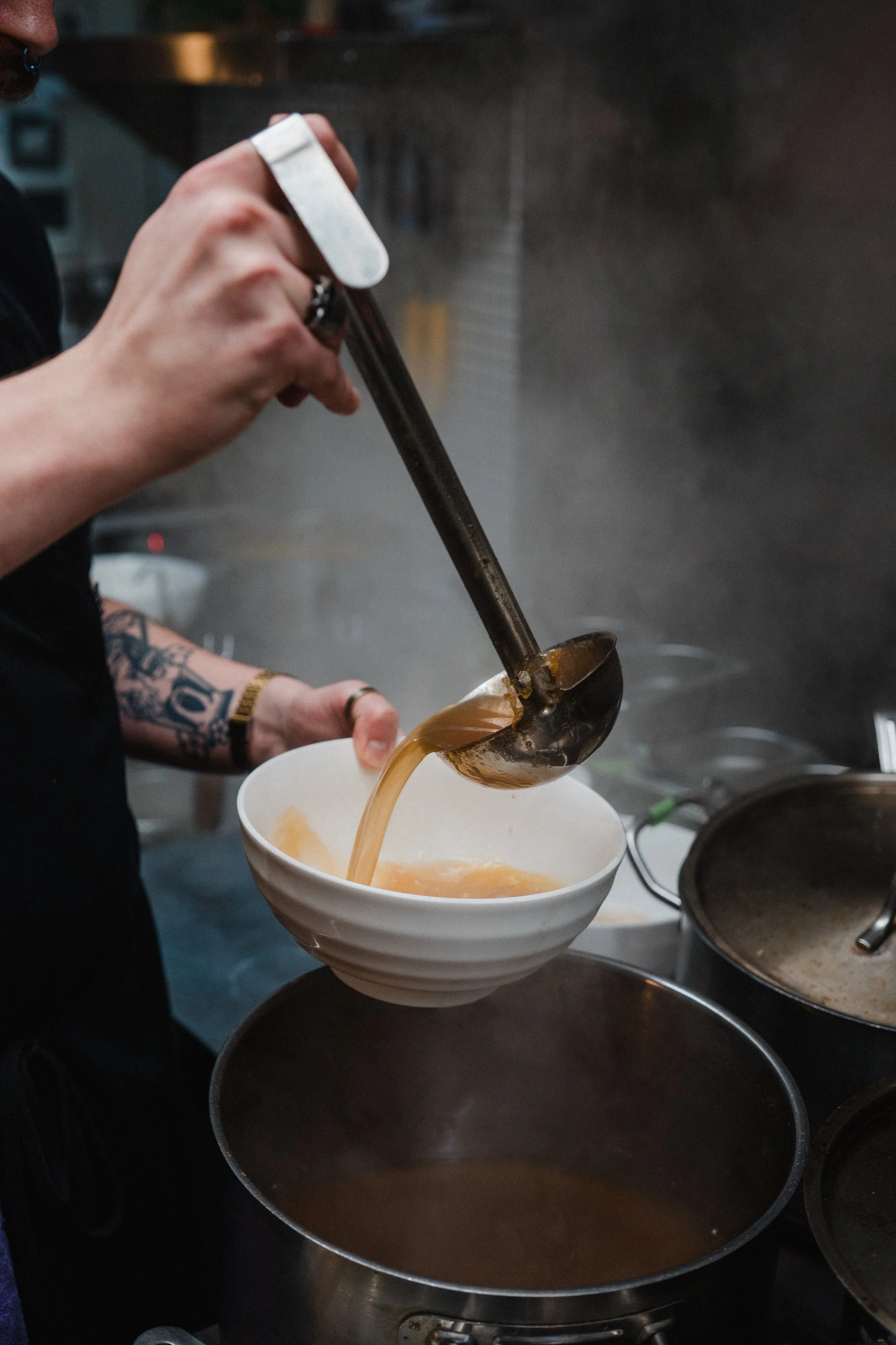 A person pours soup from a large ladle into a ramen bowl in a moody open kitchen.