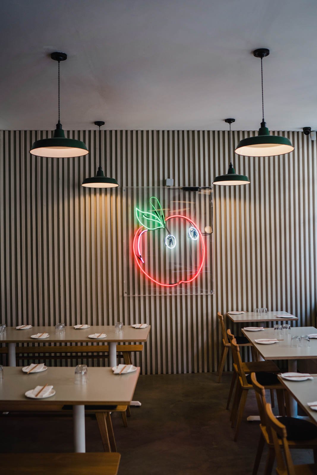 Interior of a restaurant with wood-paneled walls, four hanging green lamps, and a neon sign shaped like a cherry with green leaves and two cherries below it.