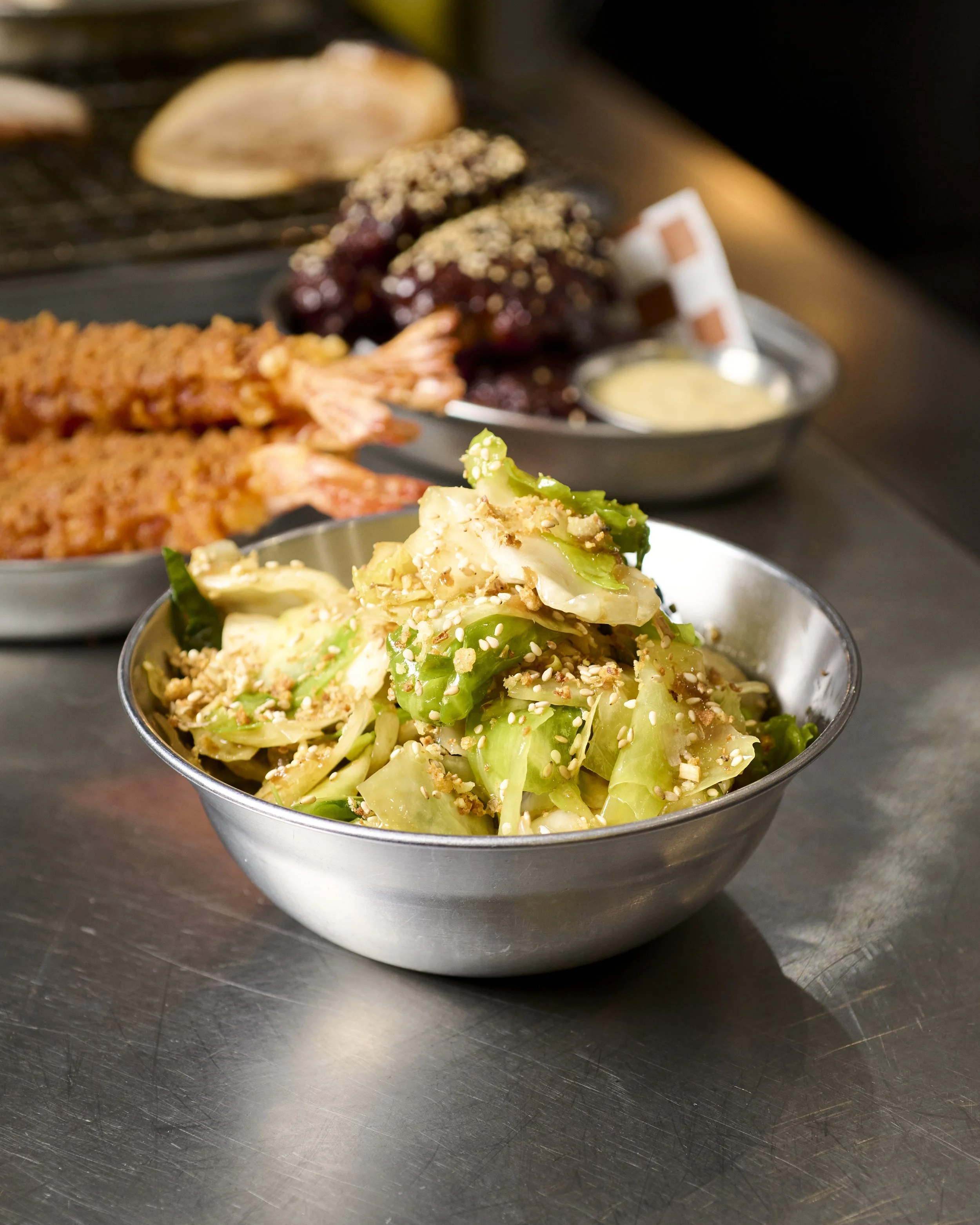 A metal bowl of chopped vegetables topped with sesame seeds in front of a blurred tray of cooked eel and meat with sauce and a shellfish in the background.
