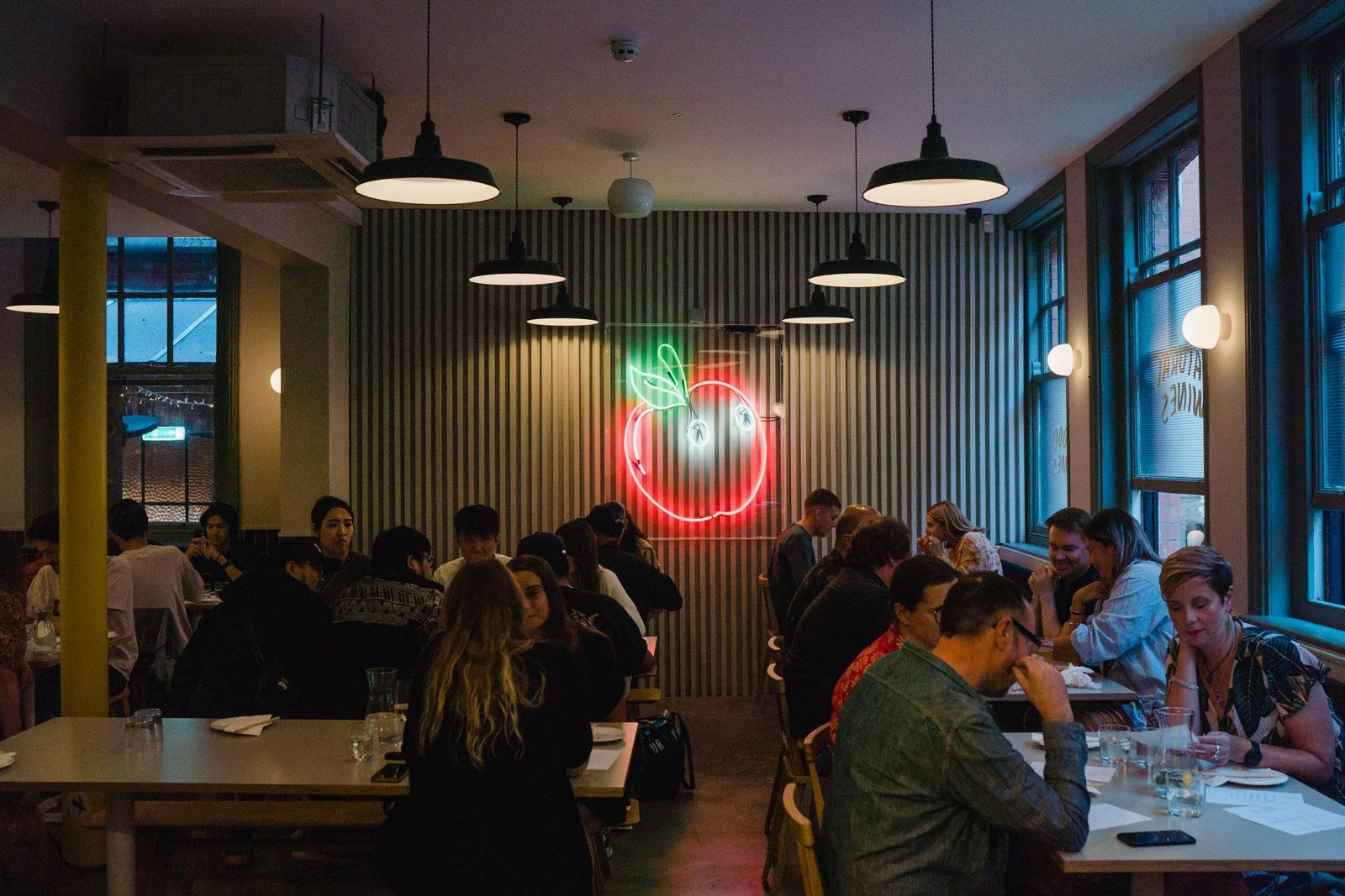 A neon sign glows on the back wall of a busy restaurant in the design of a large red apple