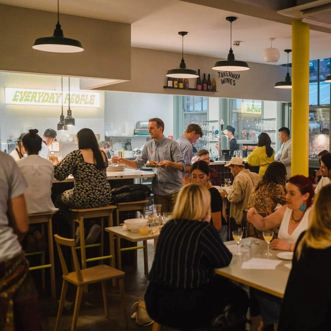 Inside Everyday People with tables, benches and counter stools full of people in a low-lit room