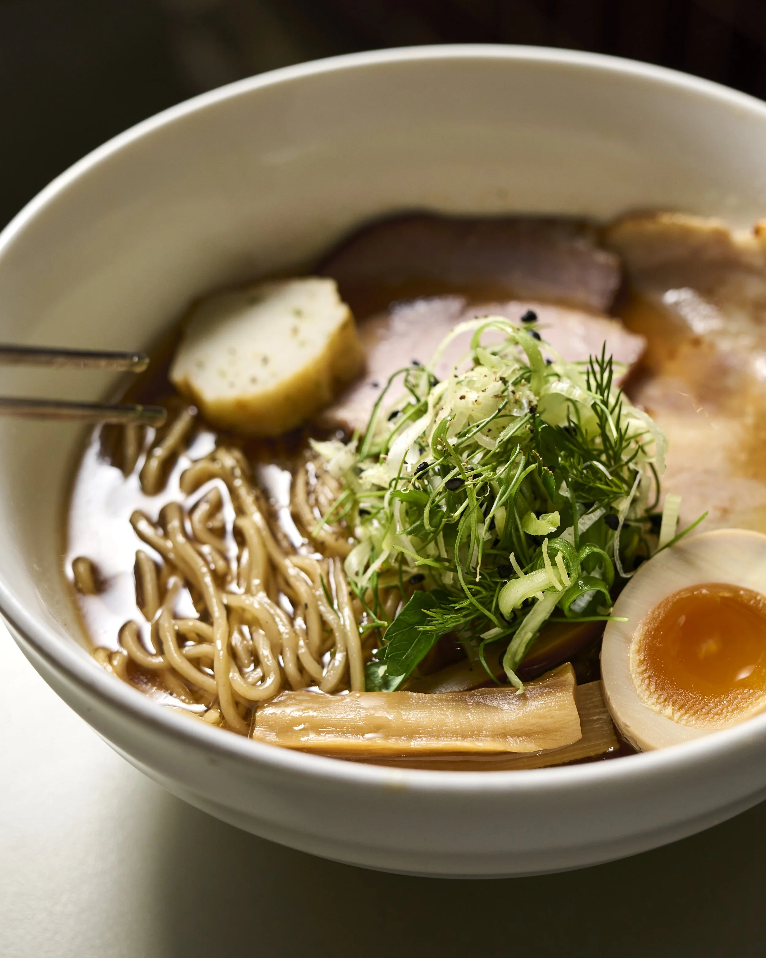 A ramen with a clear broth in a white bowl, with noodles, menma, pork coppa chashu and a jammy ramen egg
