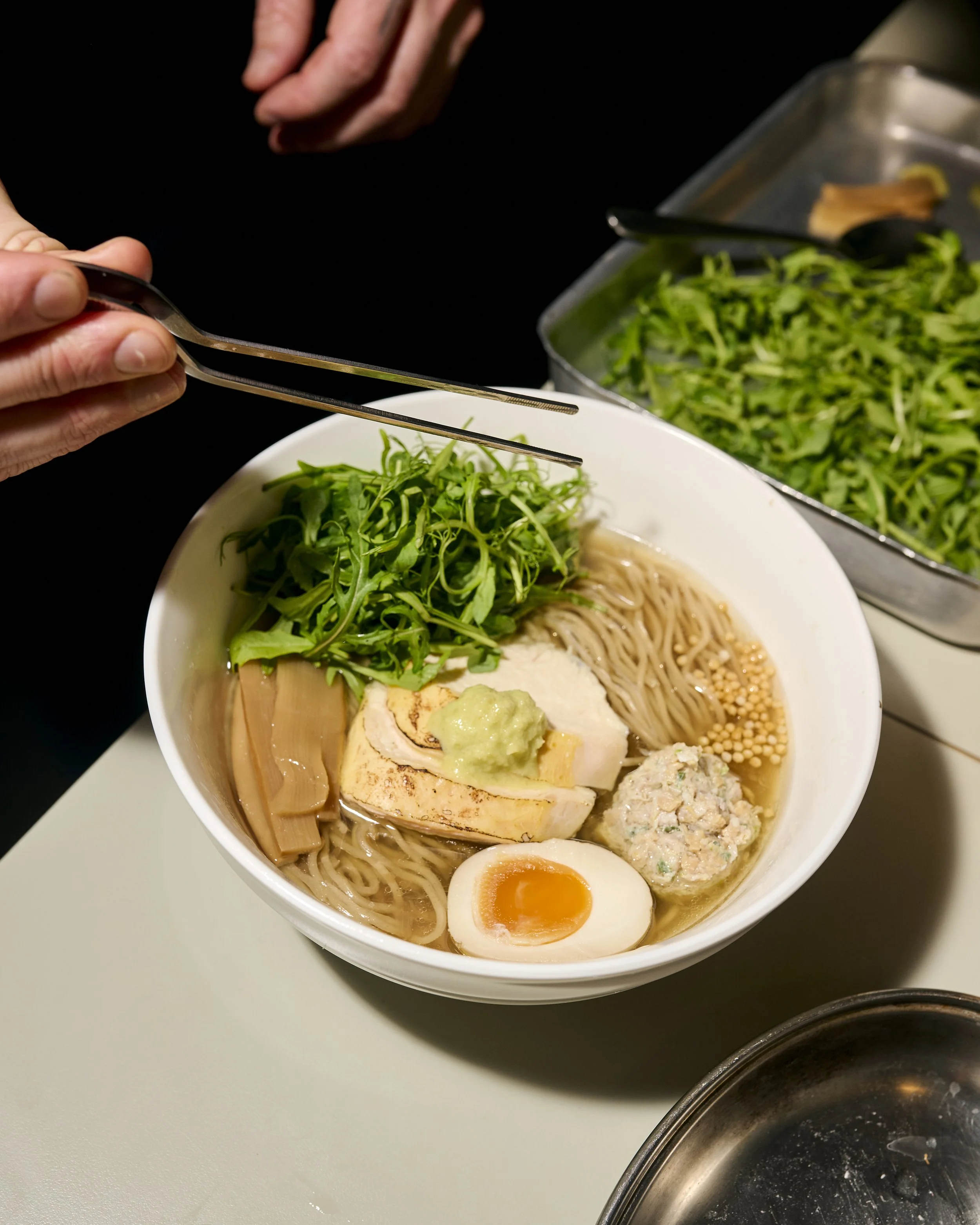 A chef is holding kitchen tweezers plating a bowl of clear-broth ramen in a white bowl