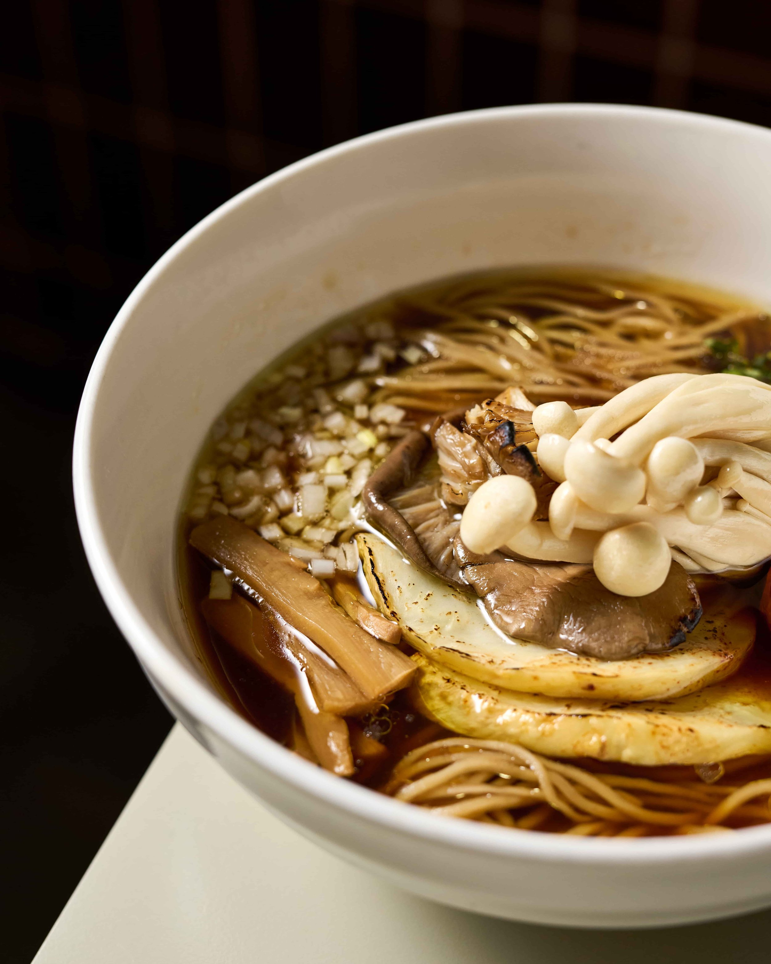 A dark but clear broth in a white bowl, with folded noodles, topped with enoki mushrooms, menma and diced shallot.
