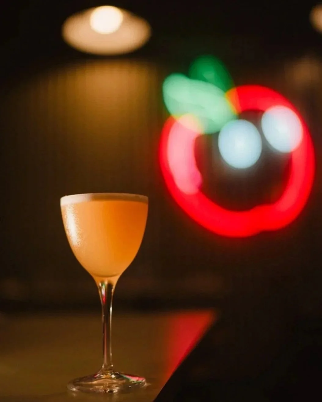 A cocktail glass with an orange-colored drink on a dark surface, with blurred colorful bokeh lights in the background.