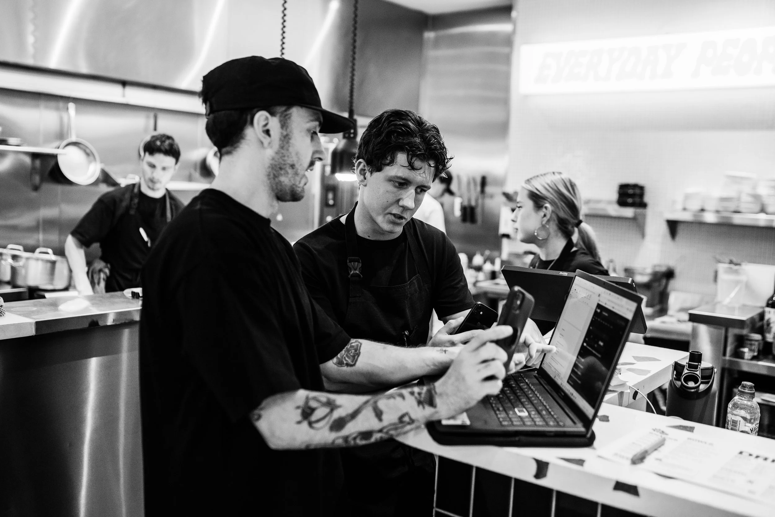 Black and white photo of two employees at a restaurant counter, both looking at a laptop. One wears a cap, and the other has wavy hair. Two other employees are in the background, one working in the open kitchen, and the other running food to tables.