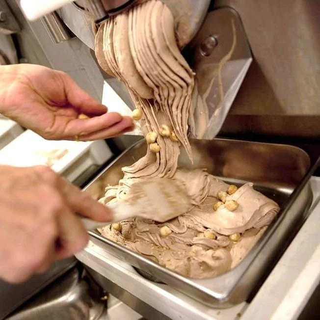 A person spreading chocolate ice cream into a metal tray, adding hazelnuts on top.