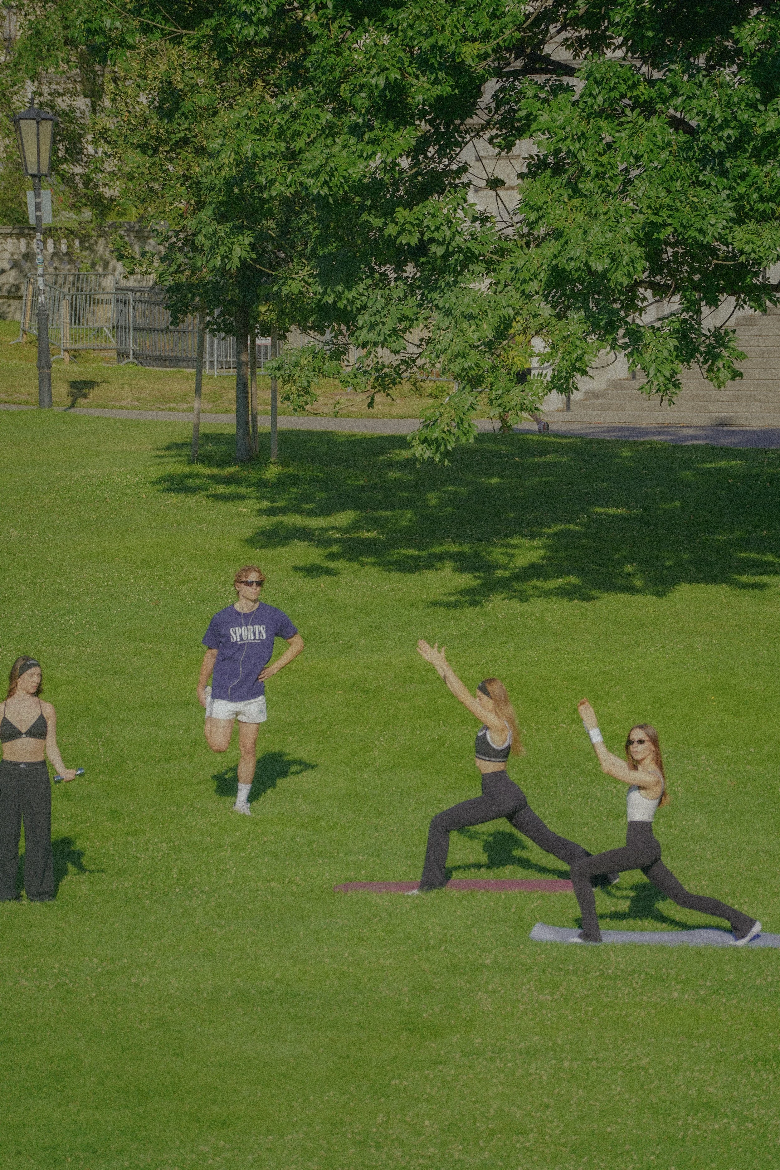 A group of people participating in an outdoor pilates class on a grassy area under a tree, with some individuals in the pilates pose lunging with their arms extended, and others standing and observing.