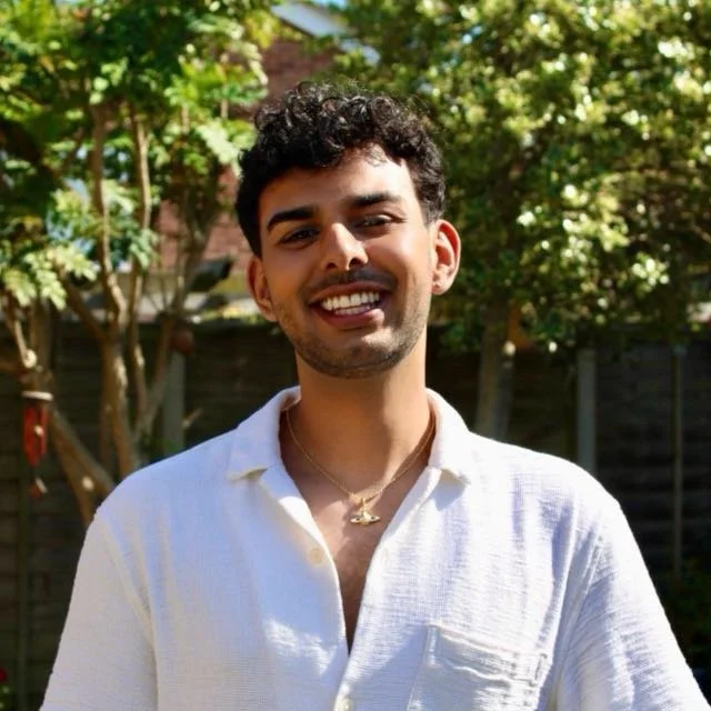Young man with curly dark hair smiling outdoors in a sunny garden, wearing a white collared shirt and a gold necklace.