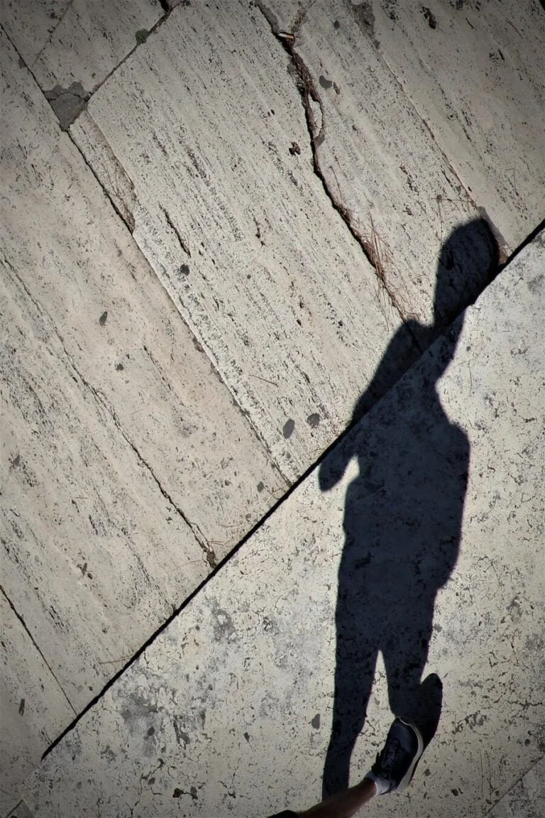 Shadow of a person with an apple-shaped head, walking on a cracked concrete sidewalk.