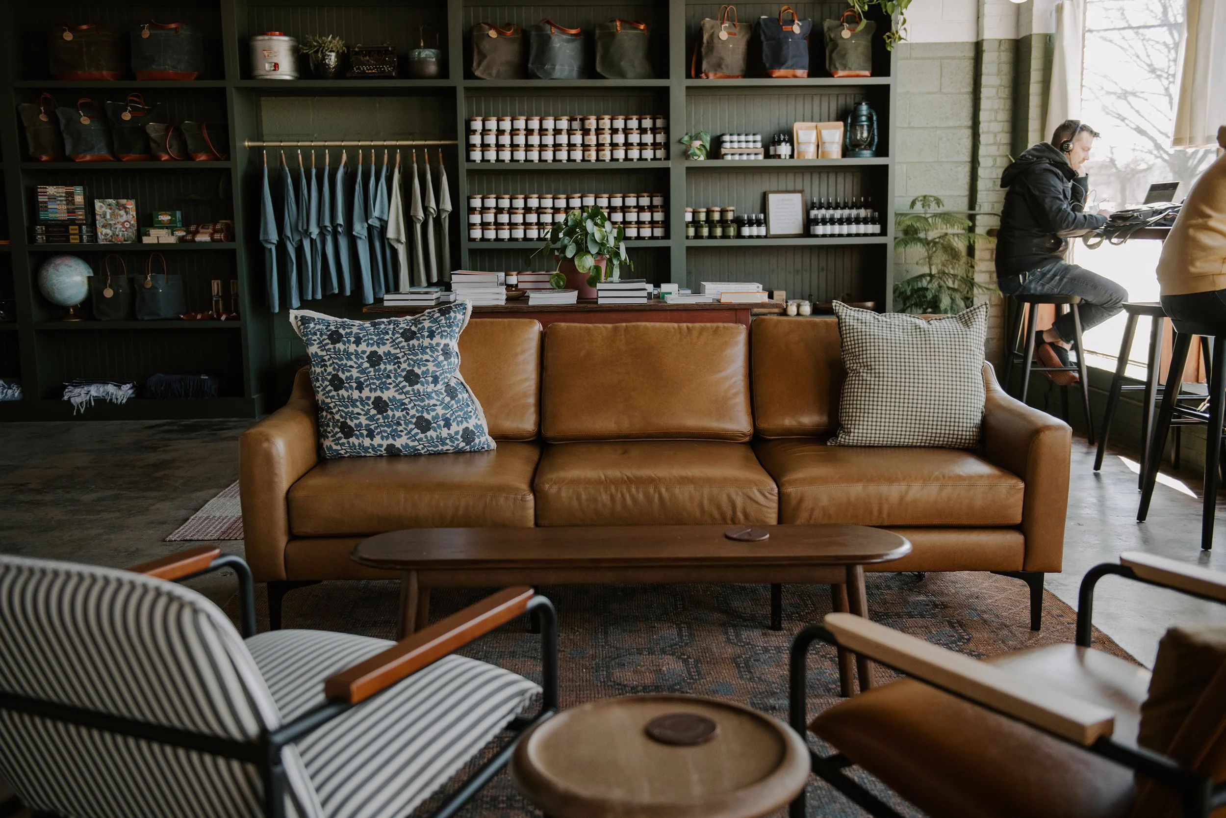 Cozy indoor space with a tan leather sofa, patterned cushions, and a wooden table in front. Behind the sofa is a dark green shelf filled with handmade goods, candles and books. Customers work at the window bar.