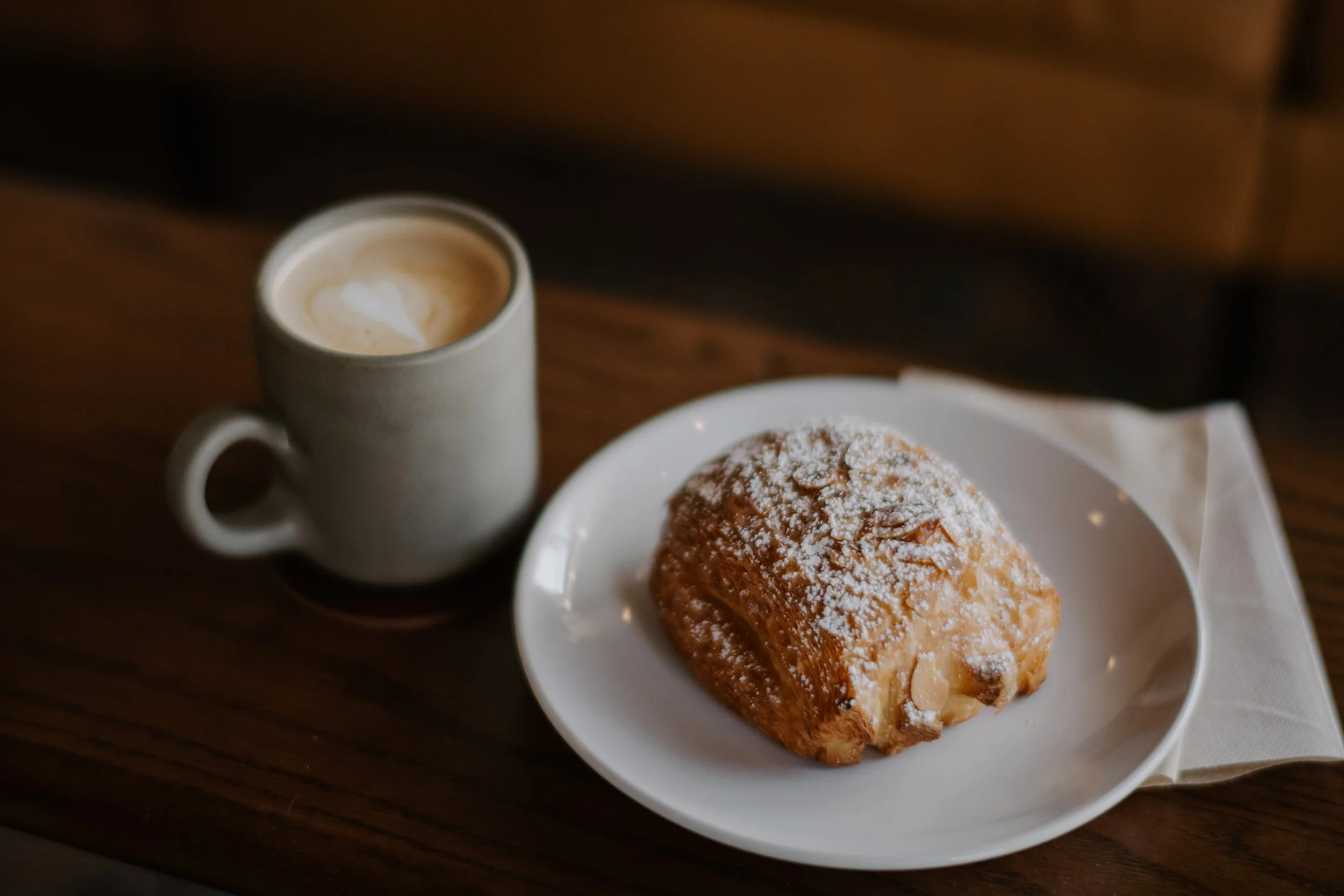 A latte with foam art next to a plate with an almond croissant dusted with powdered sugar on a wooden table.