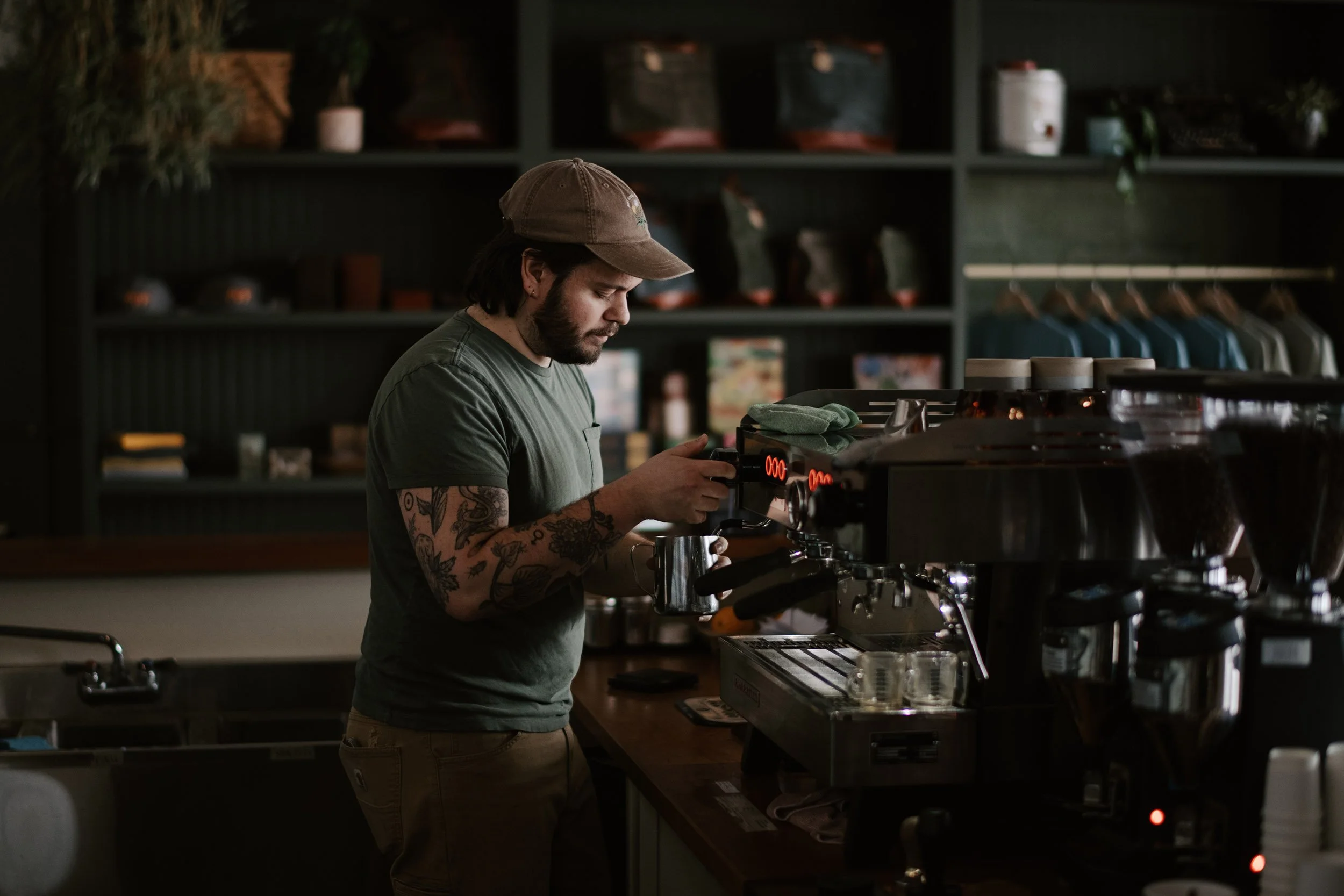 A tattoed barista making espresso in a dark, modern cafe interior with shelves of handmade products in the background.