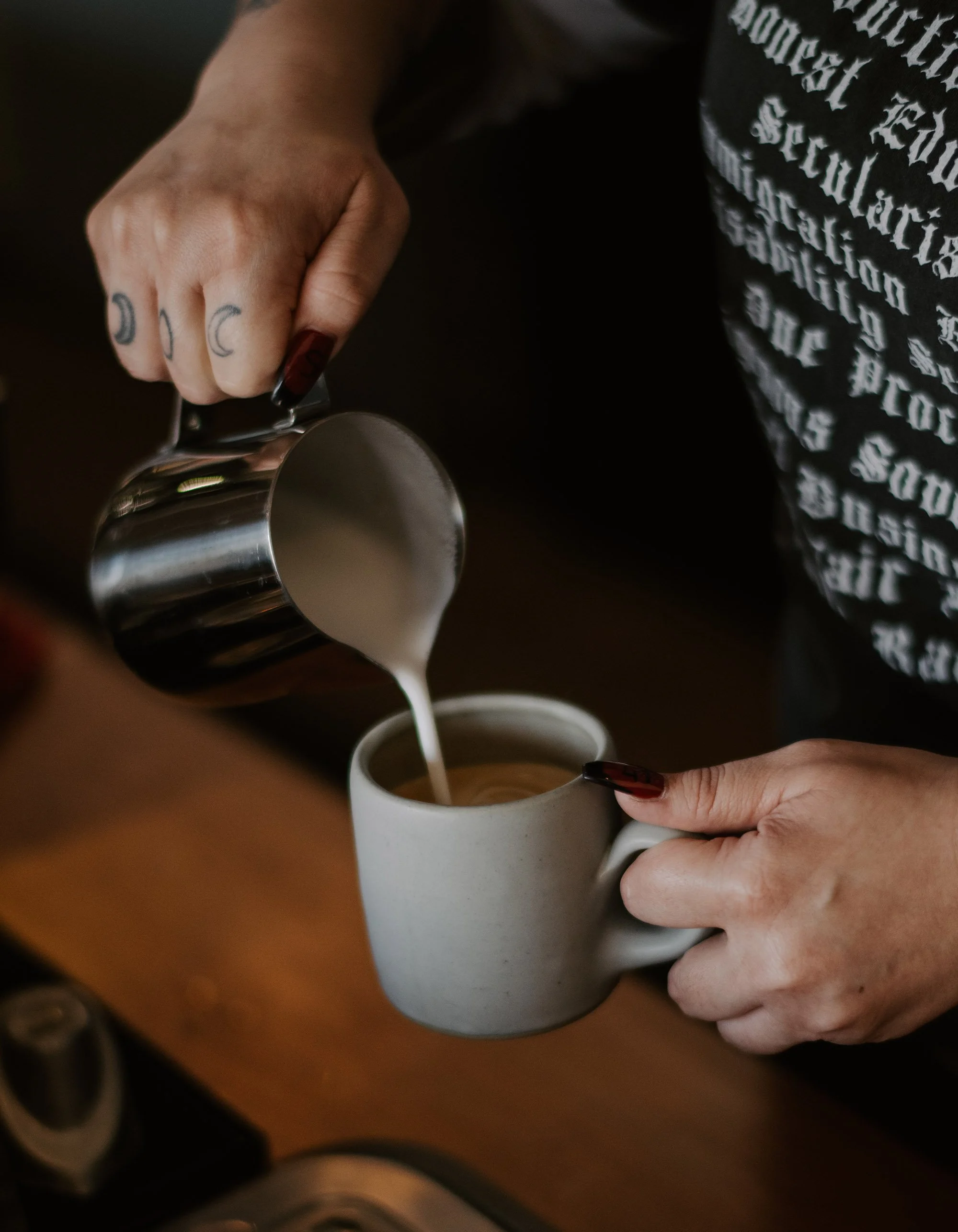 A barista's hand holding a metallic pitcher pouring steamed milk into a ceramic mug.