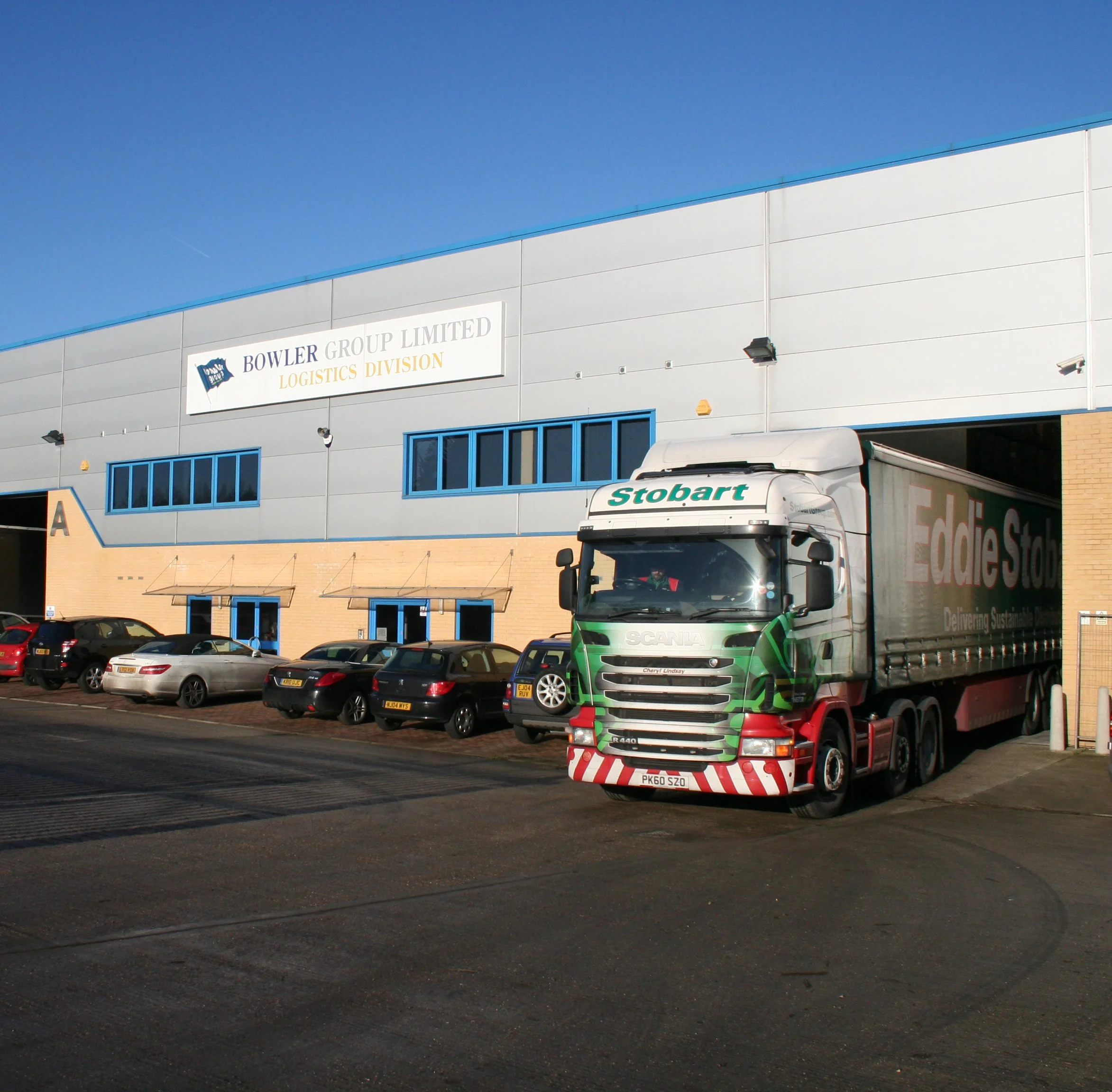 Exterior of a warehouse or industrial building with parked cars and vehicles in the parking lot, including a white truck with green accents and a trailer labeled 'Eddie Stobart'. The building has a sign for 'Bowler Group Limited Logistics Division' and features blue window frames and outdoor lighting fixtures.