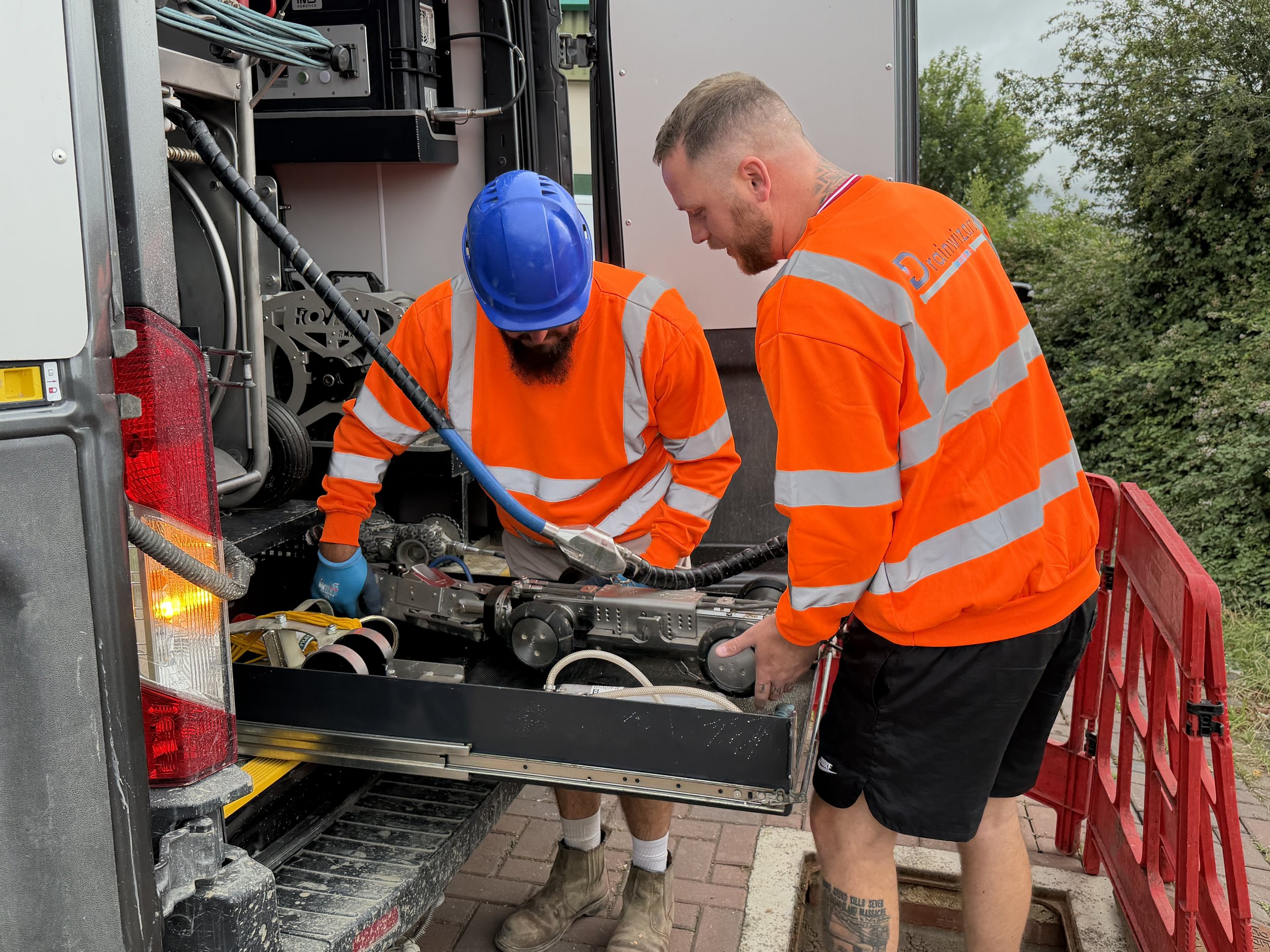 Two men wearing orange safety shirts, one with a blue hard hat, stand working on an open utility truck with electronic and mechanical equipment. They are outdoors, with trees and a cloudy sky in the background.