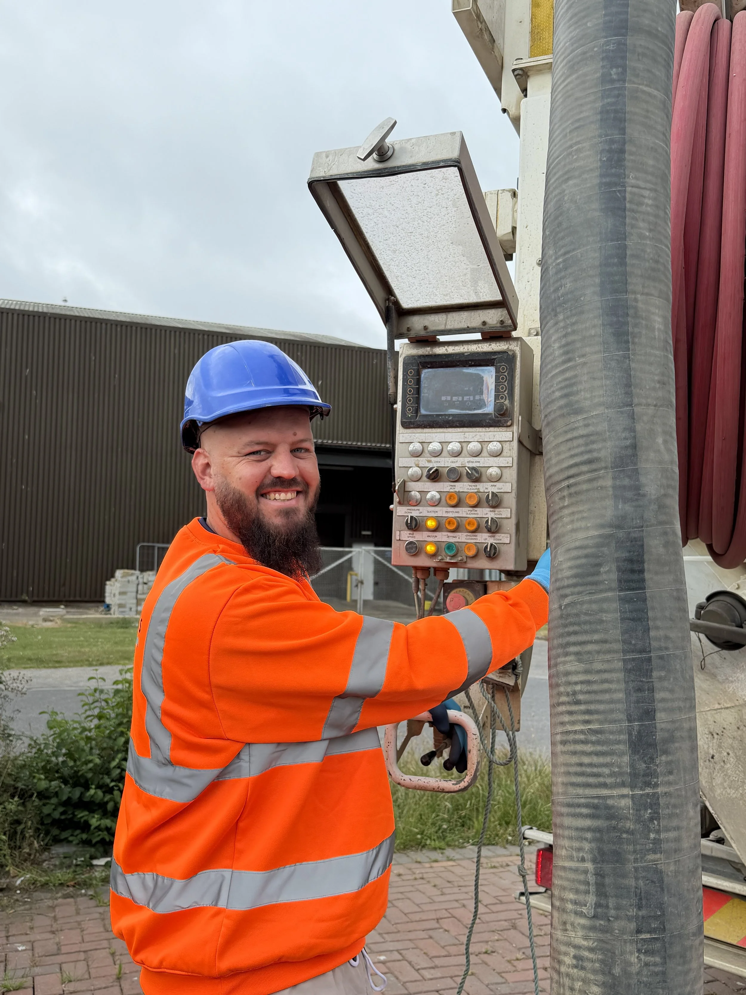 A construction worker in an orange safety vest and blue hard hat is working with electrical or control equipment, possibly at a construction site, outdoors on a cloudy day.