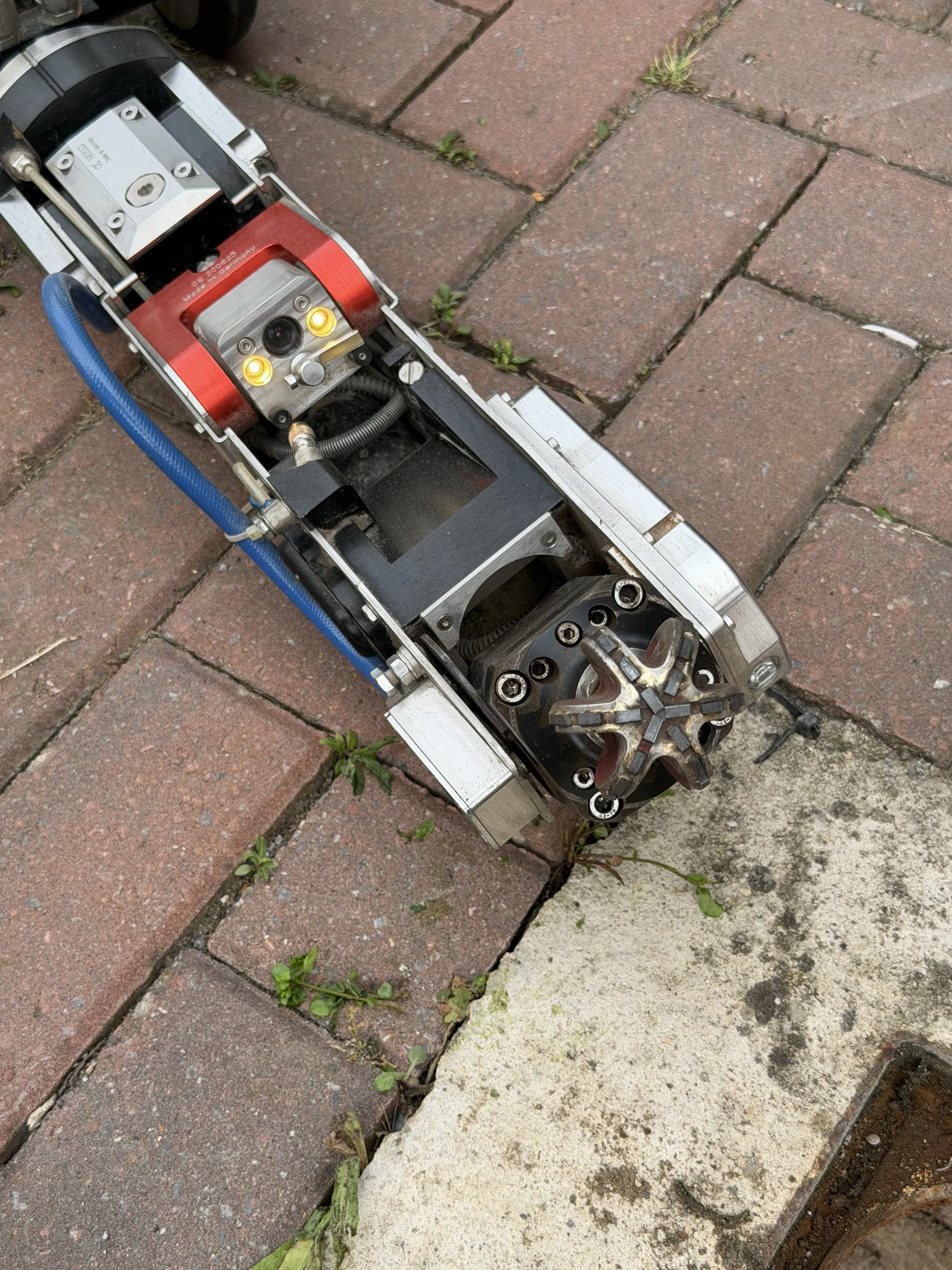 Close-up of a mechanical device or part on a brick sidewalk, with some weeds growing between the bricks.