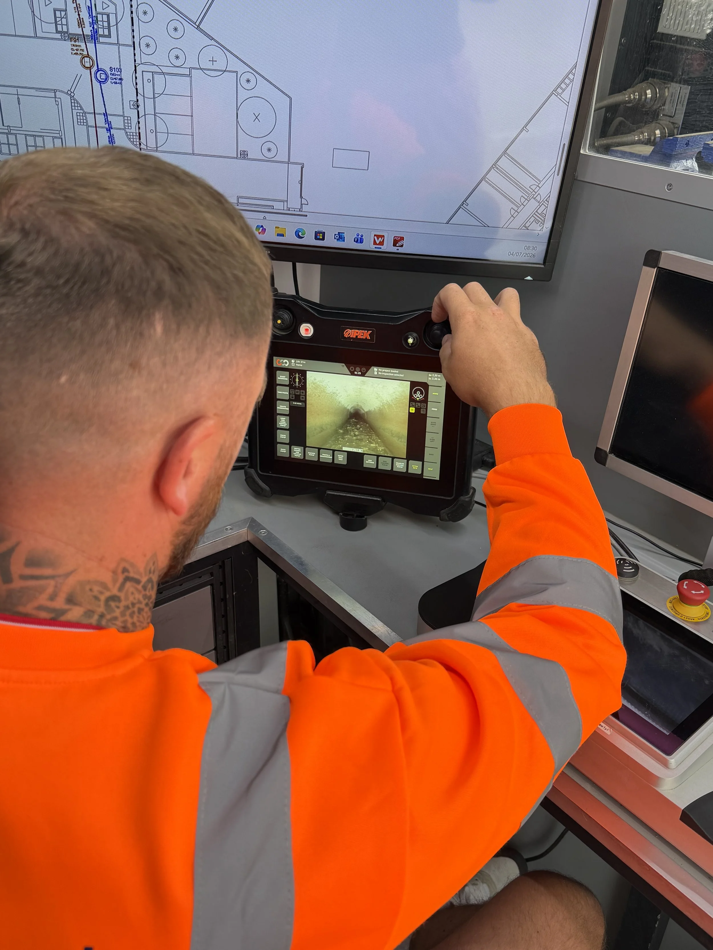 A person with short blonde hair and tattoos on their neck is operating a control panel with a small screen displaying tunnel images. They are wearing an orange safety jacket and are seated in front of a computer monitor showing a blueprint or technical drawing.