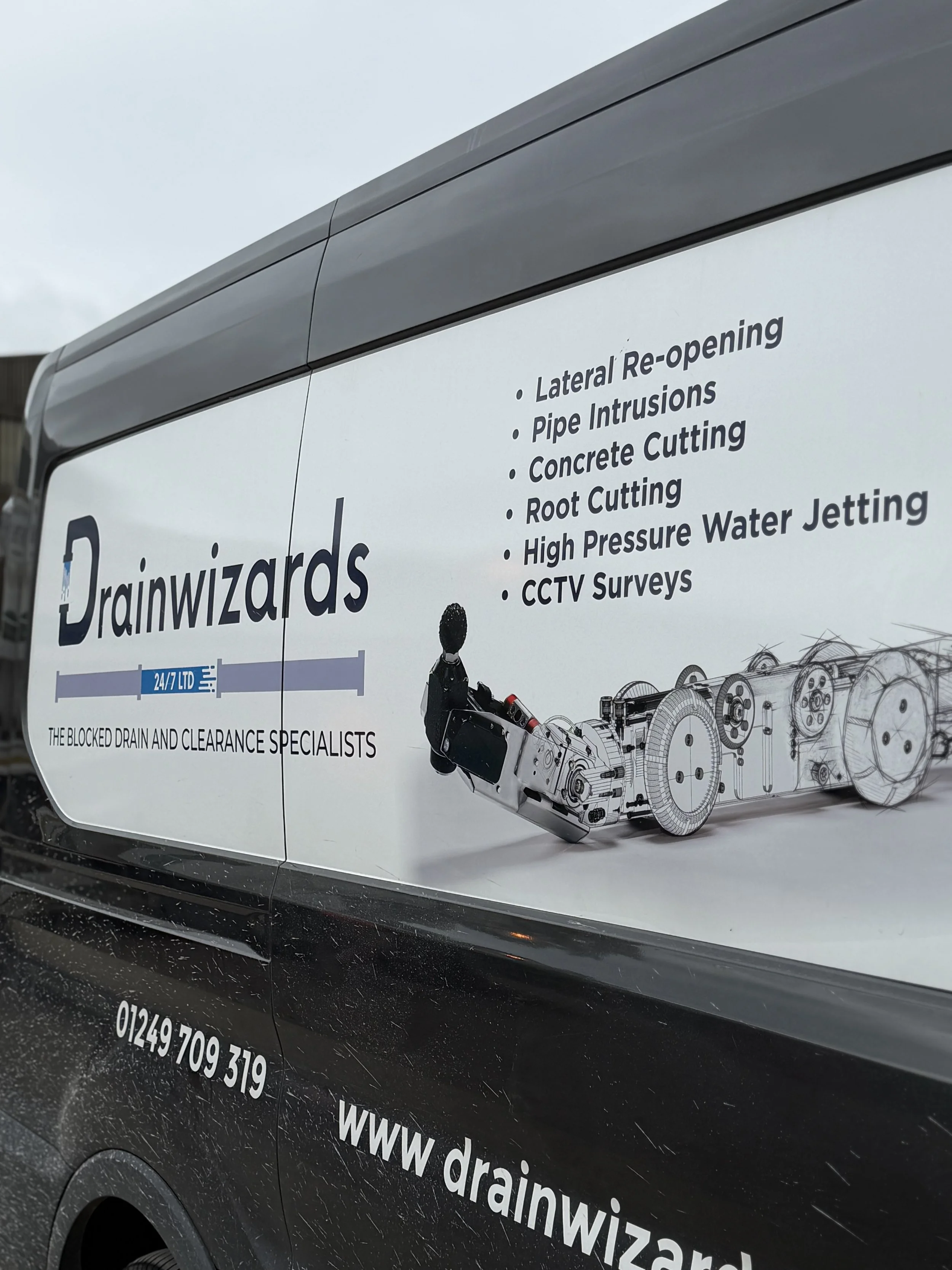 Sign on a vehicle for Drainwizards, a drain and clearance specialist company. The sign lists services including lateral re-opening, pipe intrusions, concrete cutting, root cutting, high pressure water jetting, and CCTV surveys. The sign features a technical drawing of drain components and shows part of a small person in work attire.