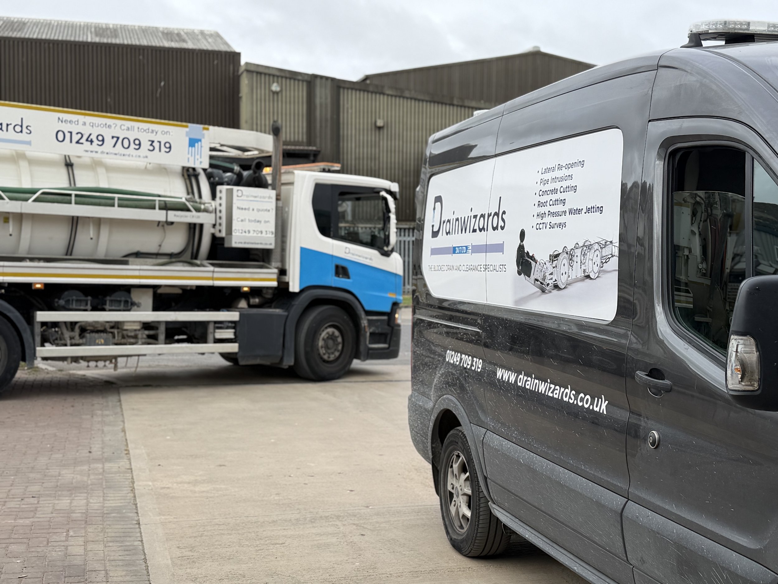 Two service vehicles parked outdoors against a backdrop of industrial buildings. One vehicle is a truck with equipment on its cargo bed, and the other is a van with branding for Drain Wizards, listing services such as pipe re-opening and concrete cutting.