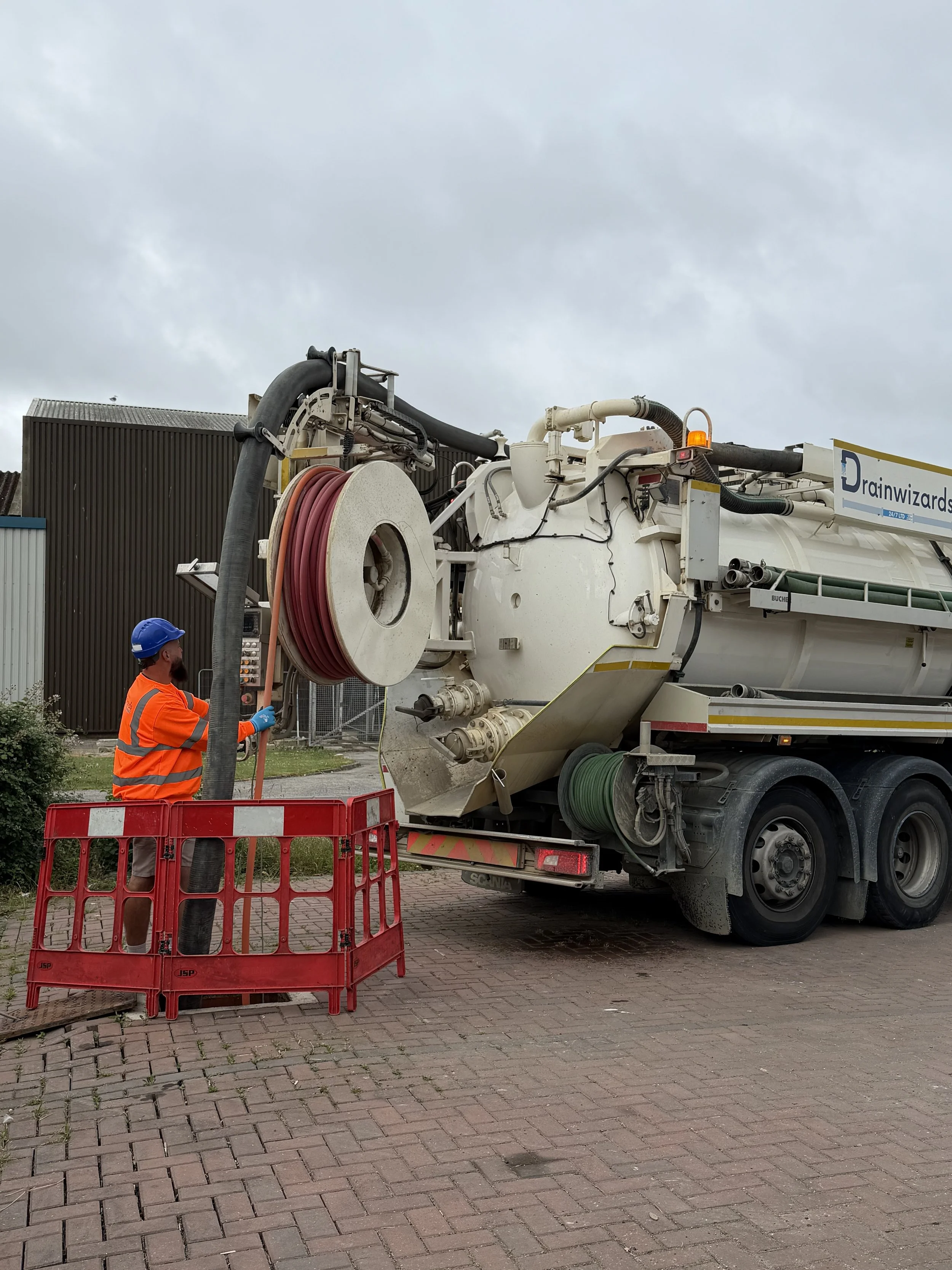 Worker in orange safety vest and blue helmet operating a large vacuum truck labeled 'Drainwizards' with hoses and equipment, parked on a brick-paved street under a cloudy sky.