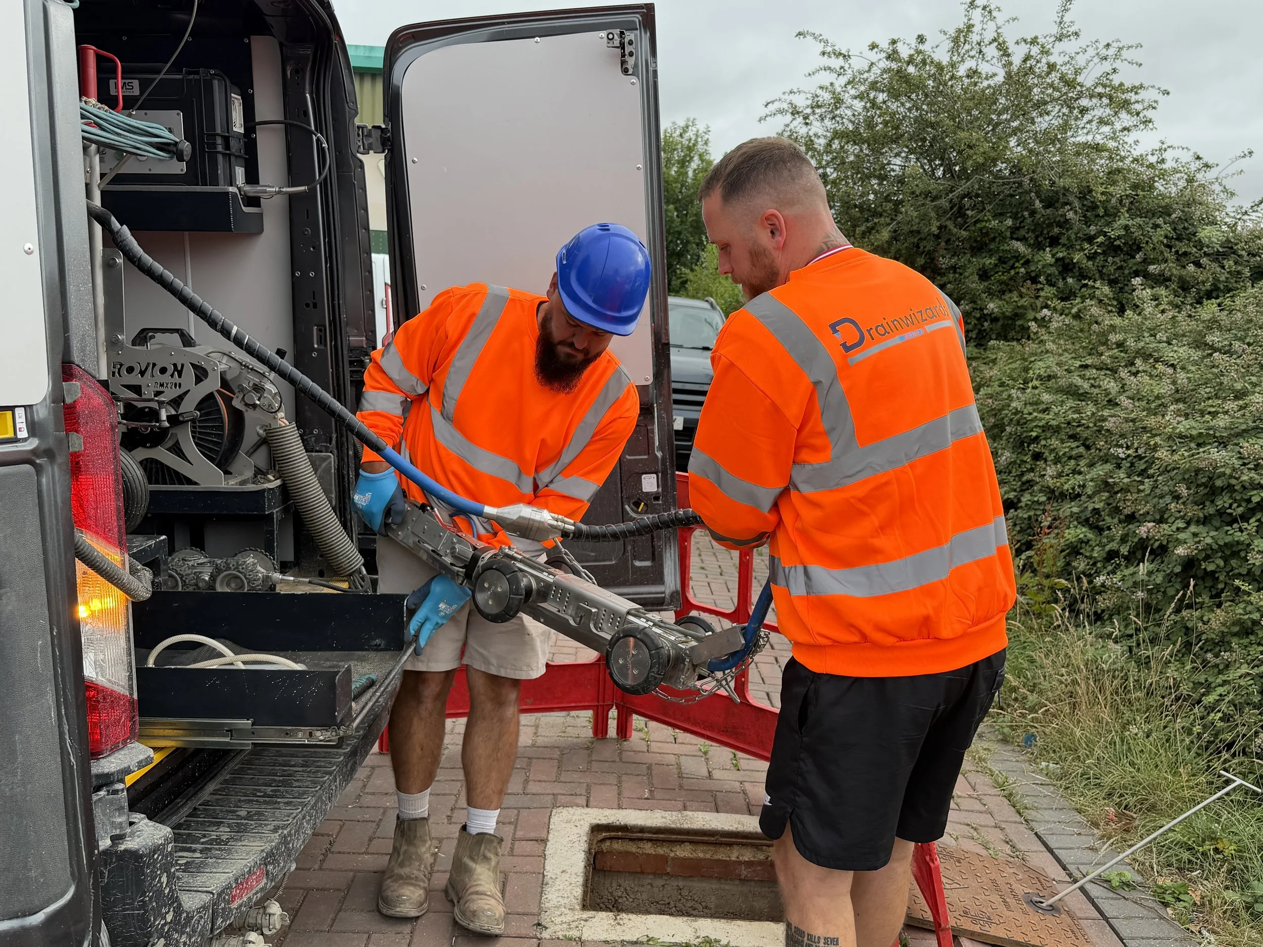 Two men wearing orange safety vests, one with a blue hard hat, working on equipment outside of a service van. One man is holding a tool while the other is assisting. They are working near an open manhole.