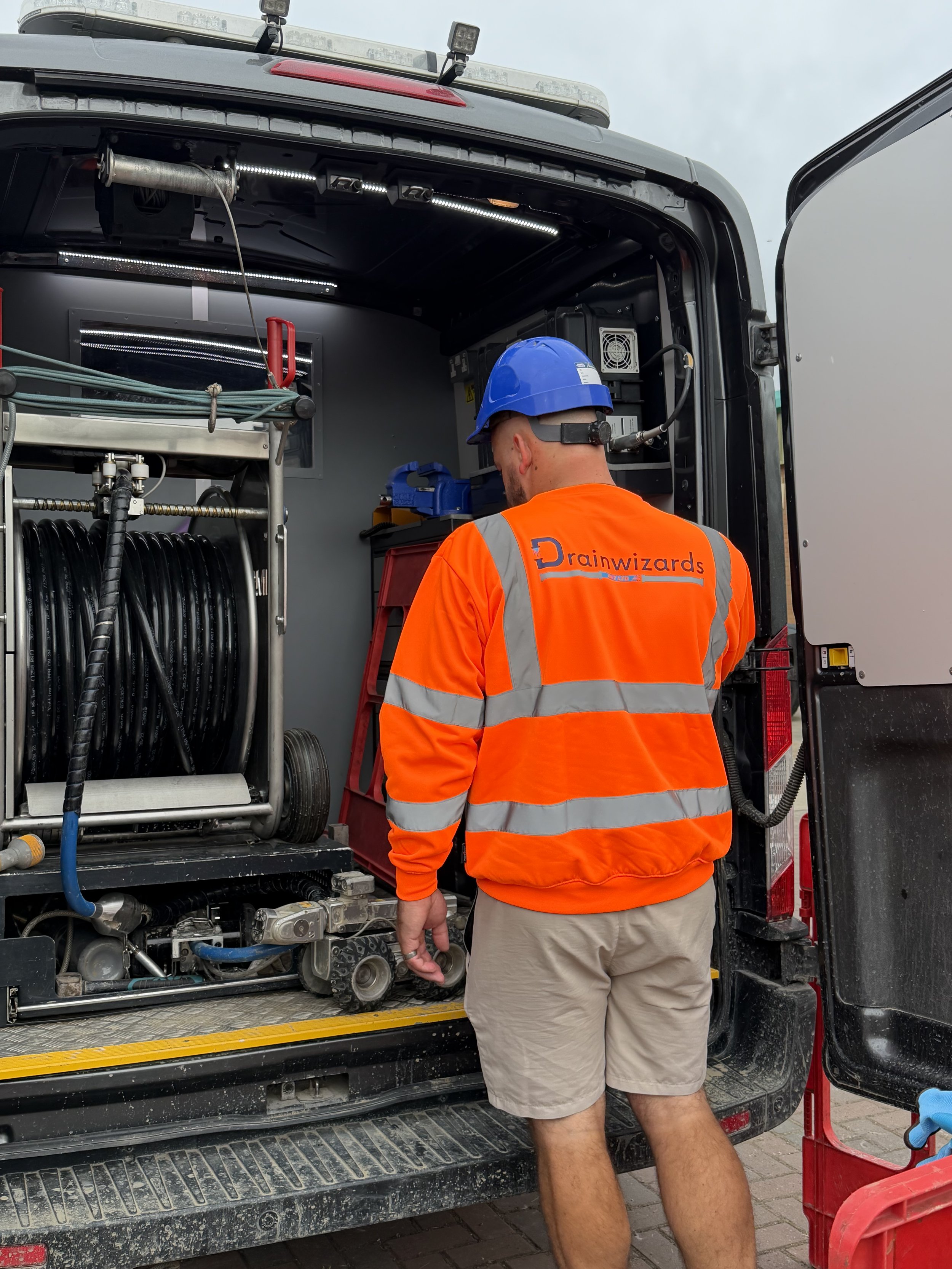 A technician wearing an orange safety vest and a blue hard hat standing next to the open compartment of a utility truck with various hoses, pipes, and tools inside. The technician is working on a piece of equipment inside the truck.