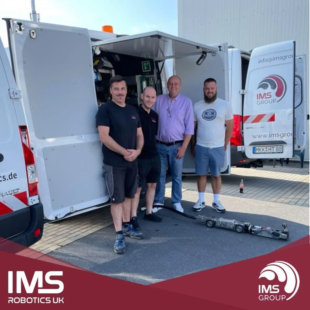 Four men standing in front of a mobile work van with the IMS Group logo, two of them wearing black shirts, one in a purple shirt, and one in a white T-shirt. The van has its side doors open, revealing equipment inside. There is a robot-like device on the ground in front of the men.