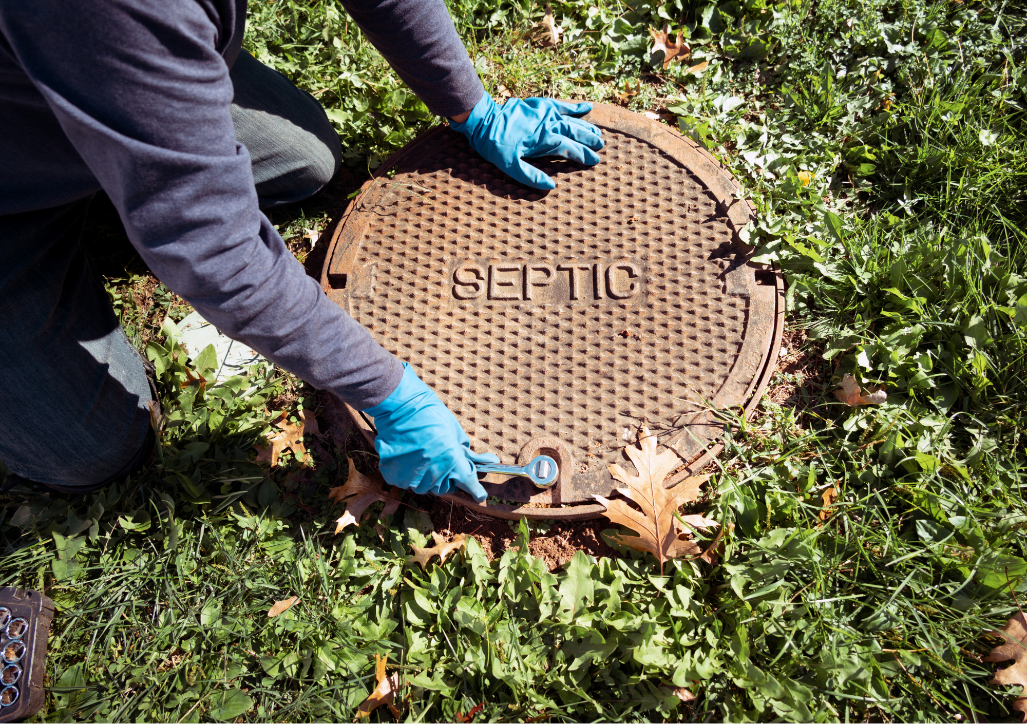 Person wearing blue gloves and a gray jacket working on a rusty septic cover in a grassy area with fallen leaves around.