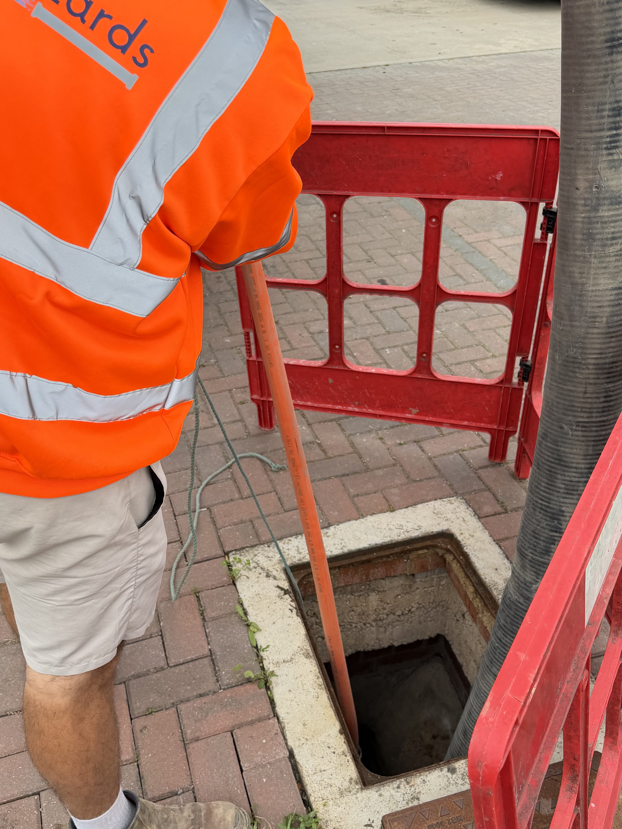 A worker wearing an orange safety vest and khaki shorts is standing next to a hole in the sidewalk. The hole appears to be a utility access point, with the cover removed. There is a red barricade next to the hole, and black hoses and cables are visible around the opening.
