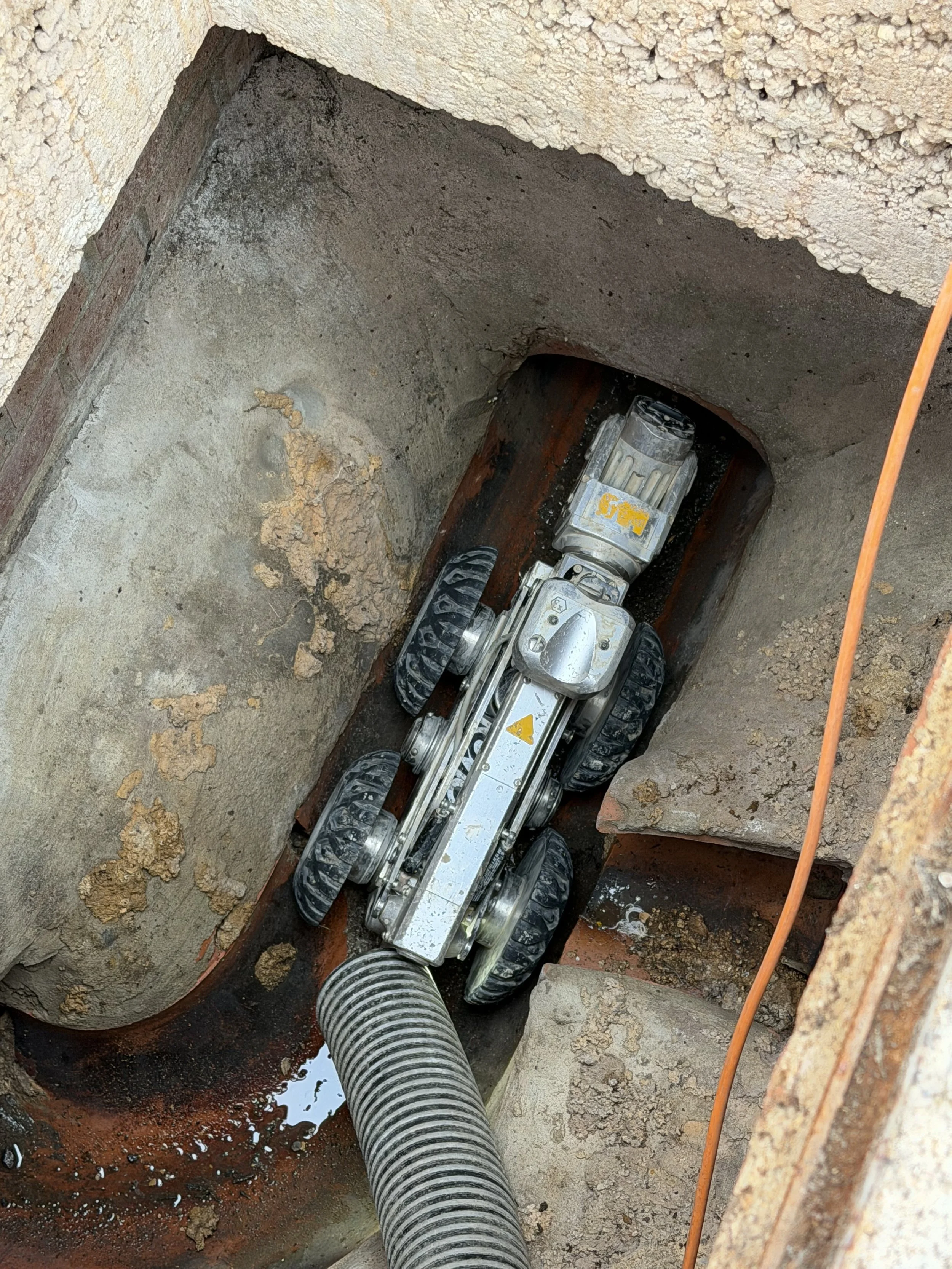 A robotic trenching machine is excavating a trench in the ground, with a large flexible pipe attached, and surrounded by dirt and concrete.