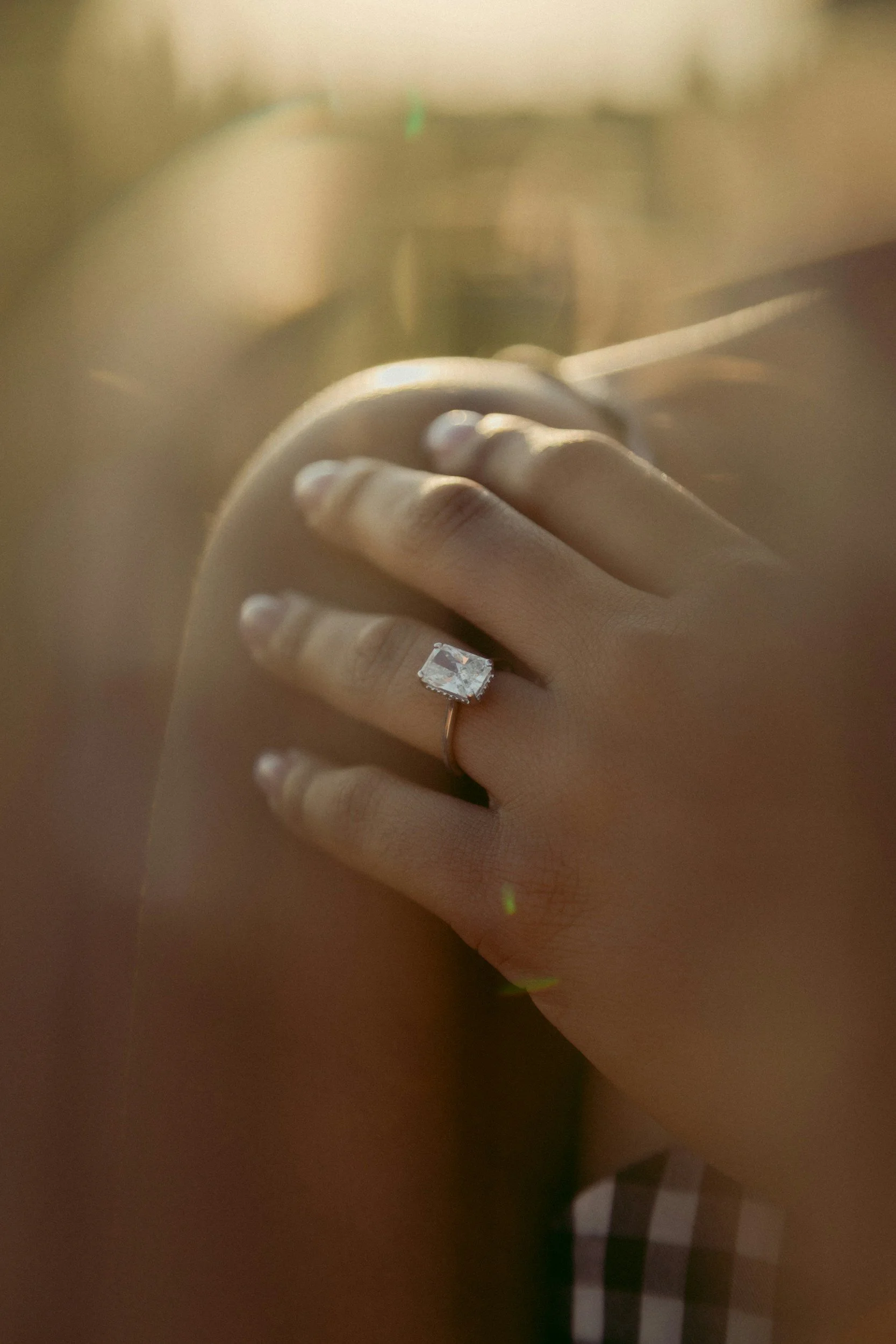 Close-up of a hand with an engagement ring on the ring finger. The background is blurred, showing a mirror and a room with warm lighting.